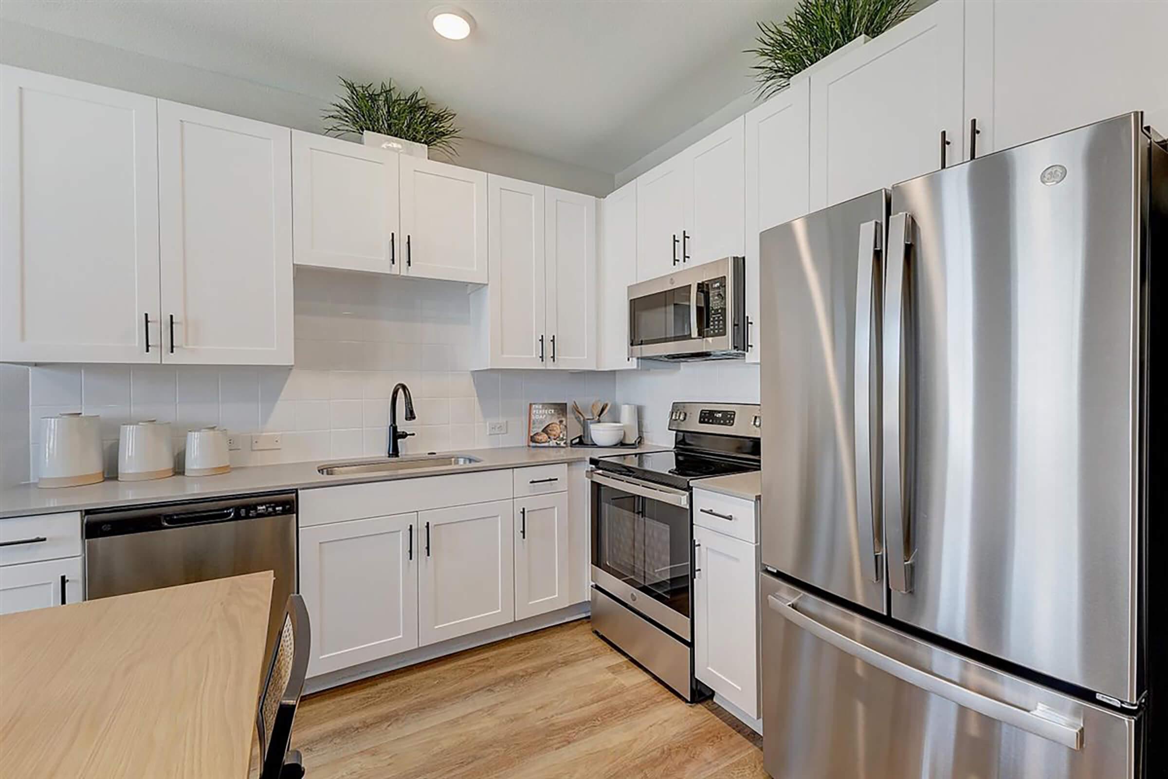 Modern kitchen with white cabinetry, stainless steel appliances including a refrigerator and oven, a sink with faucet, and a dining table with a chair. Decorative plants are placed on top of the cabinets. The flooring is light wood, contributing to a bright and airy atmosphere.