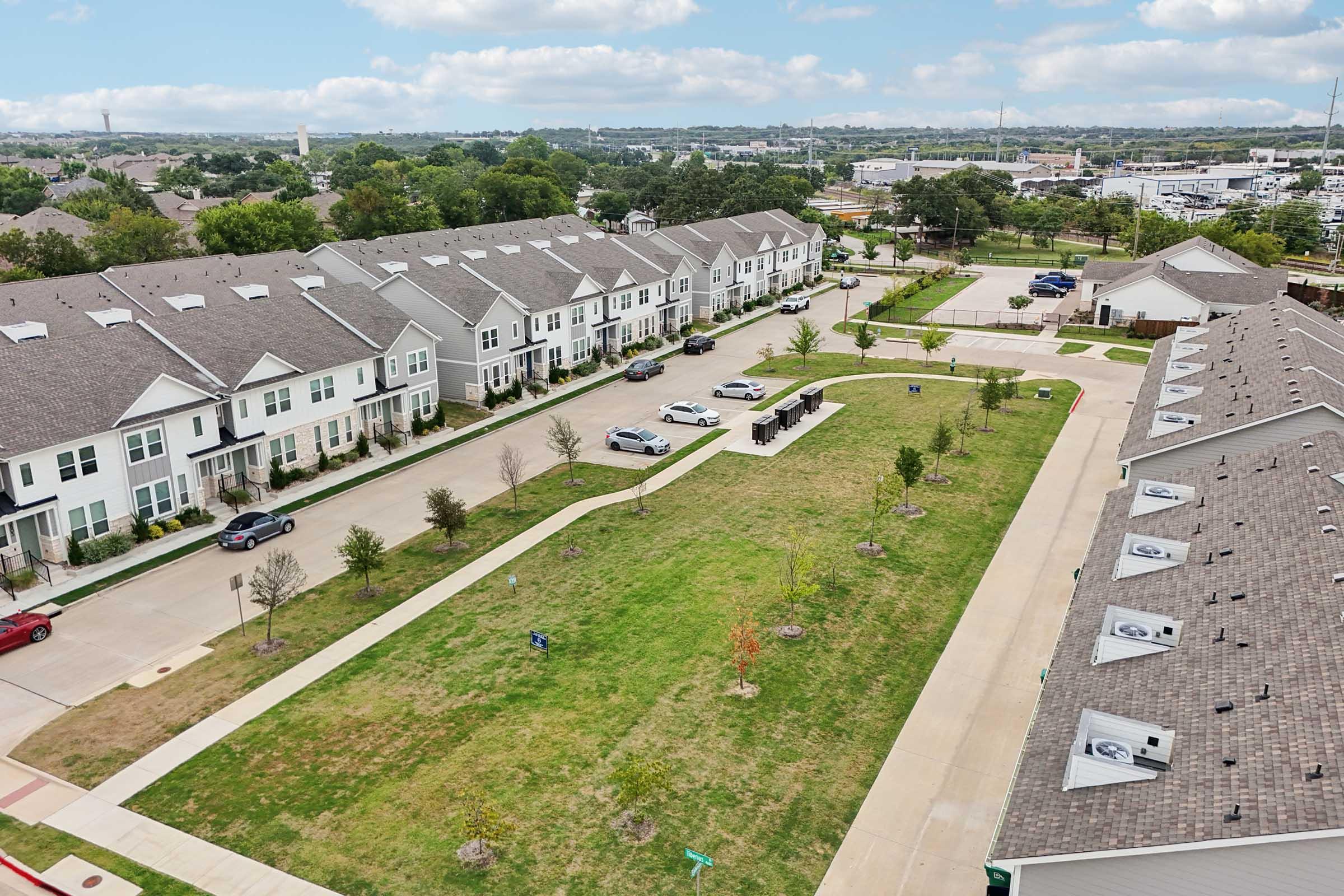Aerial view of a residential neighborhood featuring multiple row houses with gray roofs, a neatly landscaped grassy area, and a winding pathway. Several parked cars are visible along the streets, and the background shows a mix of greenery and urban development under a partly cloudy sky.