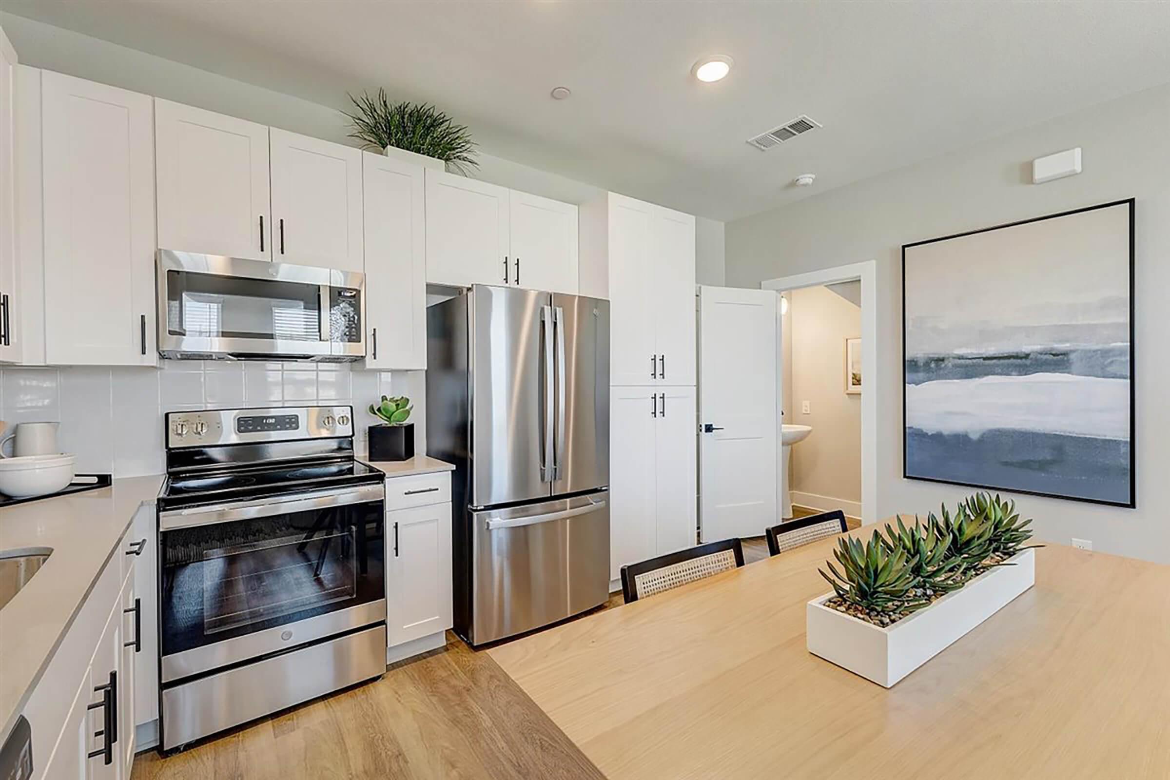 Modern kitchen featuring stainless steel appliances, including an oven and a refrigerator. Bright cabinetry with a light countertop. A dining table with succulents as a centerpiece sits in the foreground. The walls are painted in soft colors, and there's a large artwork piece on the wall in the background.