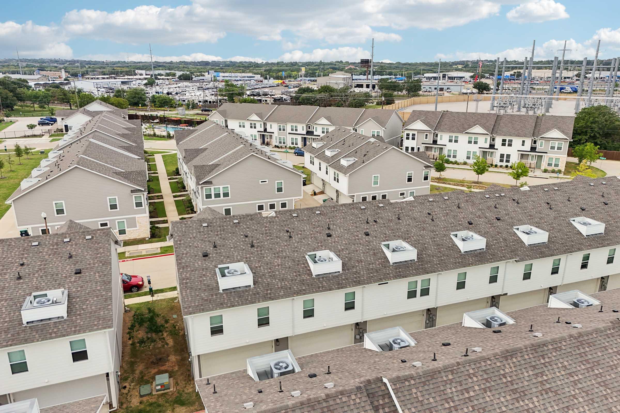 Aerial view of a residential neighborhood featuring several two-story townhouses with gray siding and sloped roofs. The foreground shows rooftops with air conditioning units, while the background includes a parking lot and distant commercial buildings under a partly cloudy sky.