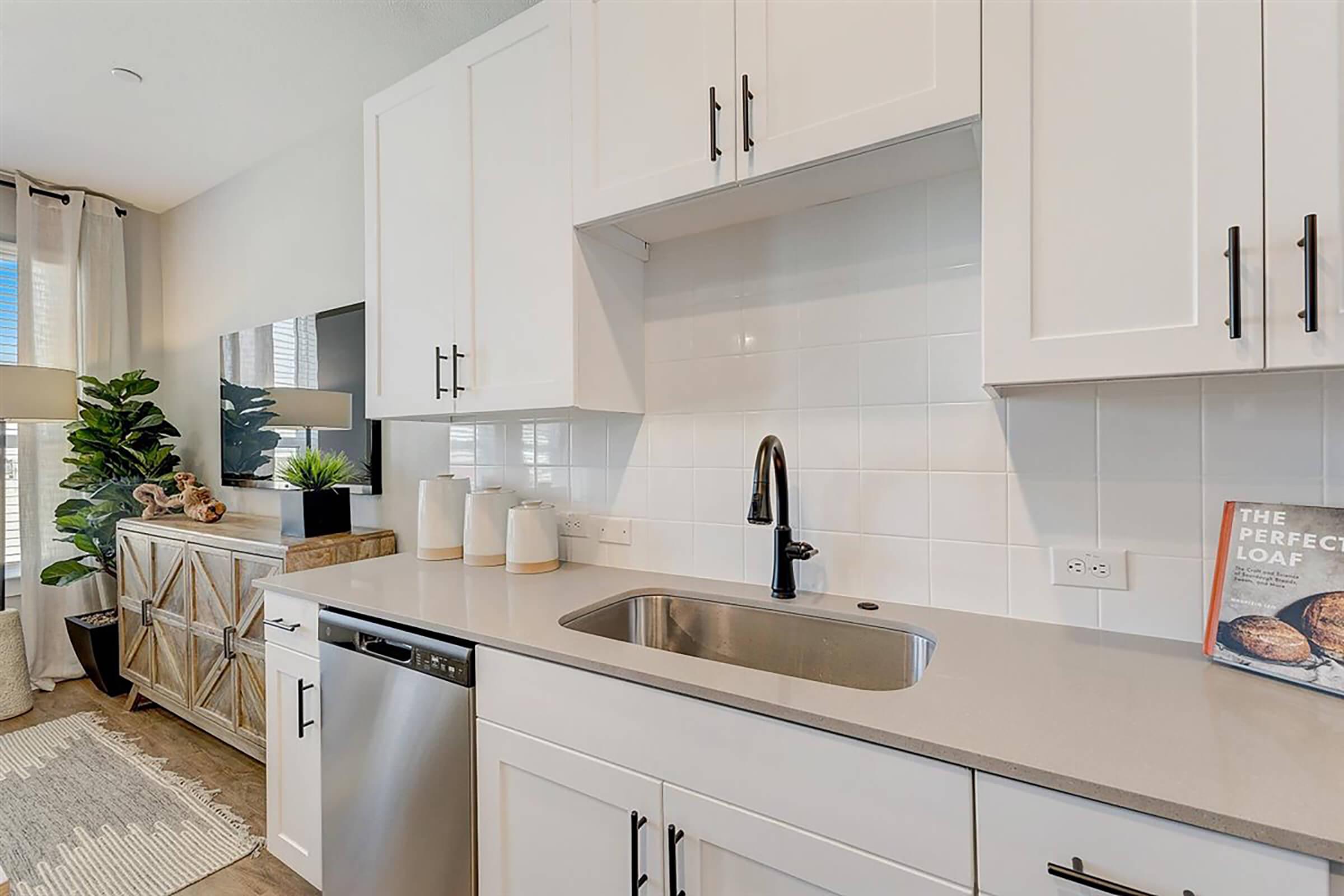 Modern kitchen with white cabinetry and a stainless steel dishwasher. A sleek black faucet is mounted above a stainless steel sink. Decor includes a green plant, decorative items on the countertop, and a book titled "The Perfect Loaf" resting against a white backsplash. Natural light filters through a window.