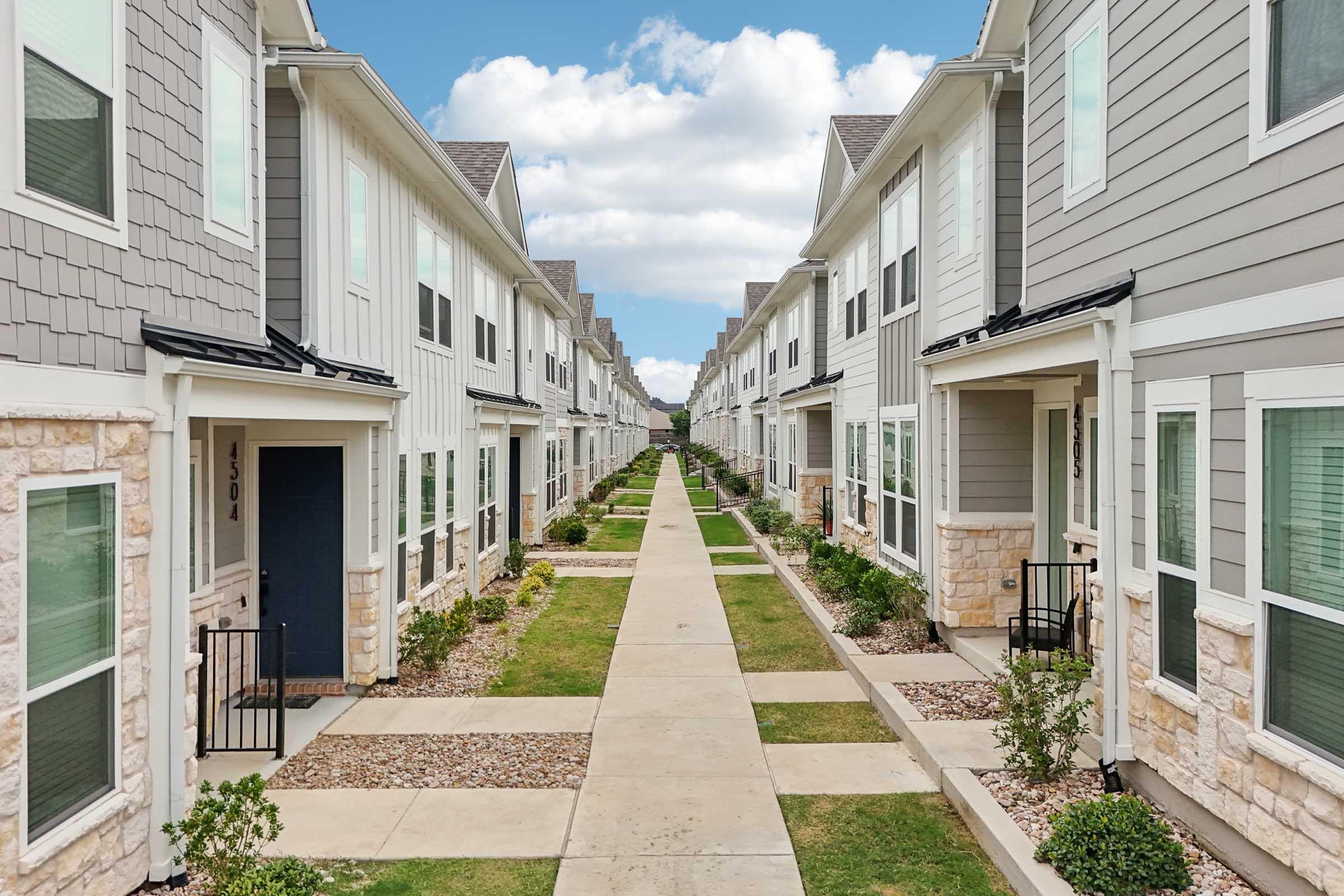 A row of modern townhouses with stone and siding exteriors, featuring landscaped pathways and neatly trimmed grass. The sky is bright with scattered clouds, creating a welcoming atmosphere in a suburban setting. Each unit has a small porch and well-defined entrance.