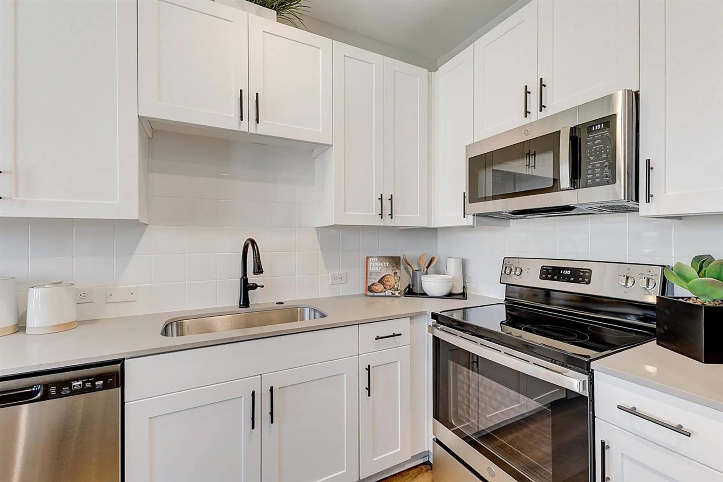 Modern kitchen featuring white cabinetry, stainless steel appliances, including a microwave, oven, and dishwasher. The countertop is sleek with a sink and a decorative plant in a black pot. Bright, airy atmosphere with a neutral color palette and minimalistic design.
