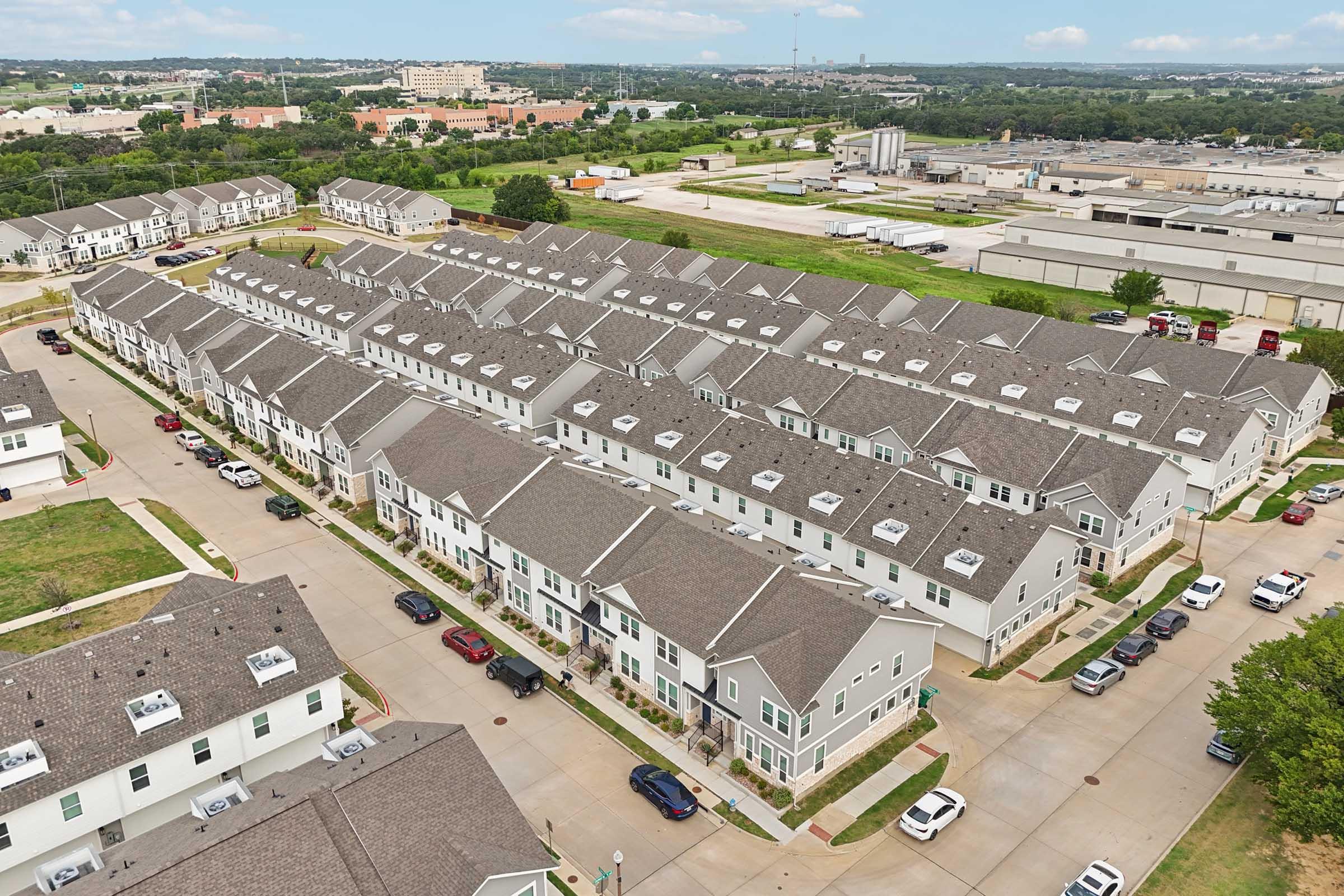 Aerial view of a residential area featuring multiple multi-family homes arranged in rows, with cars parked along the driveways. The landscape includes greenery and a mix of urban structures in the background, illustrating a suburban community setting.