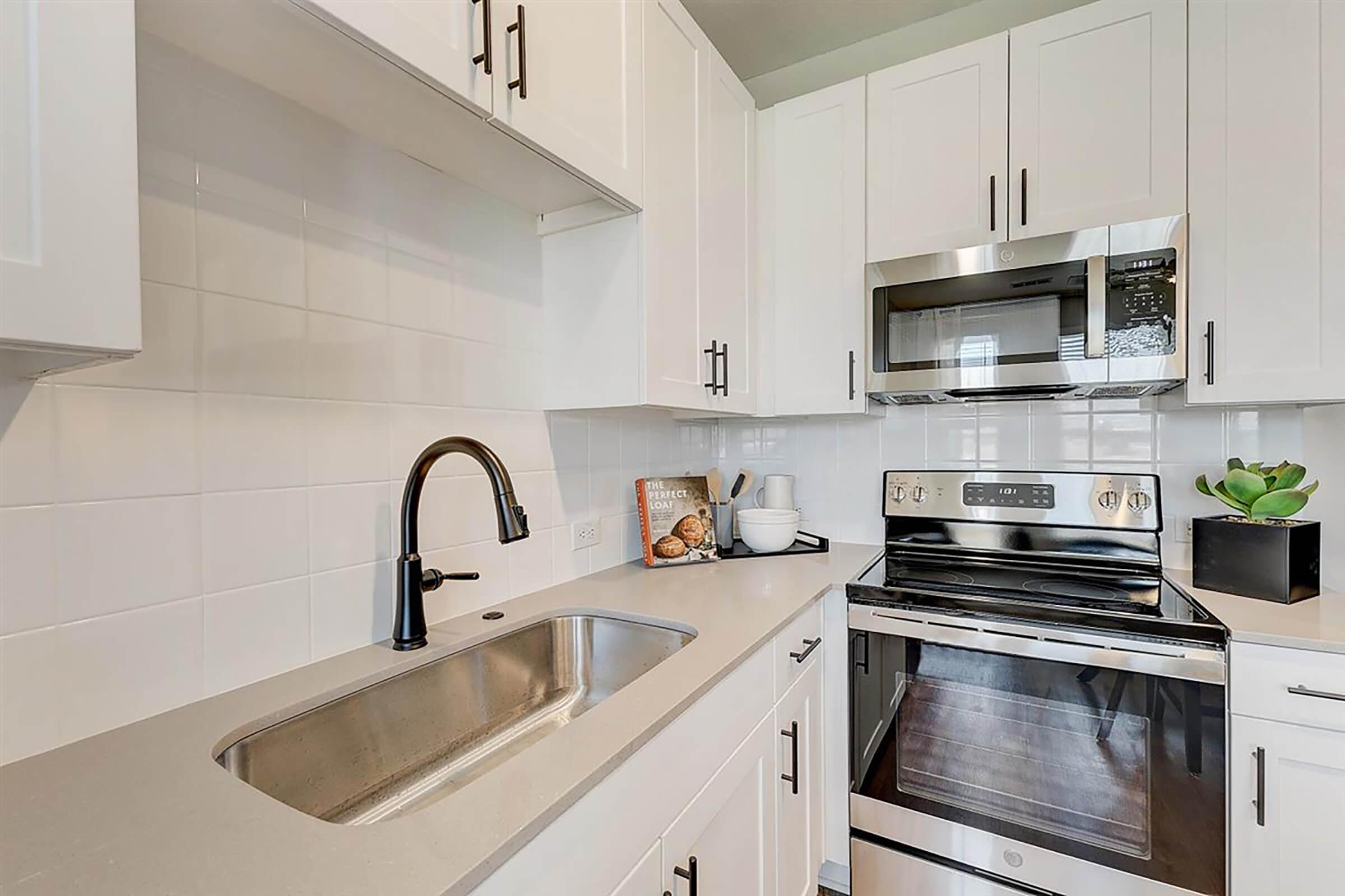 Modern kitchen featuring white cabinets, a stainless steel sink with a sleek black faucet, and an electric stove with an oven below. A microwave is mounted above the stove. A small succulent plant and kitchen utensils are displayed on the countertop, along with a cookbook.