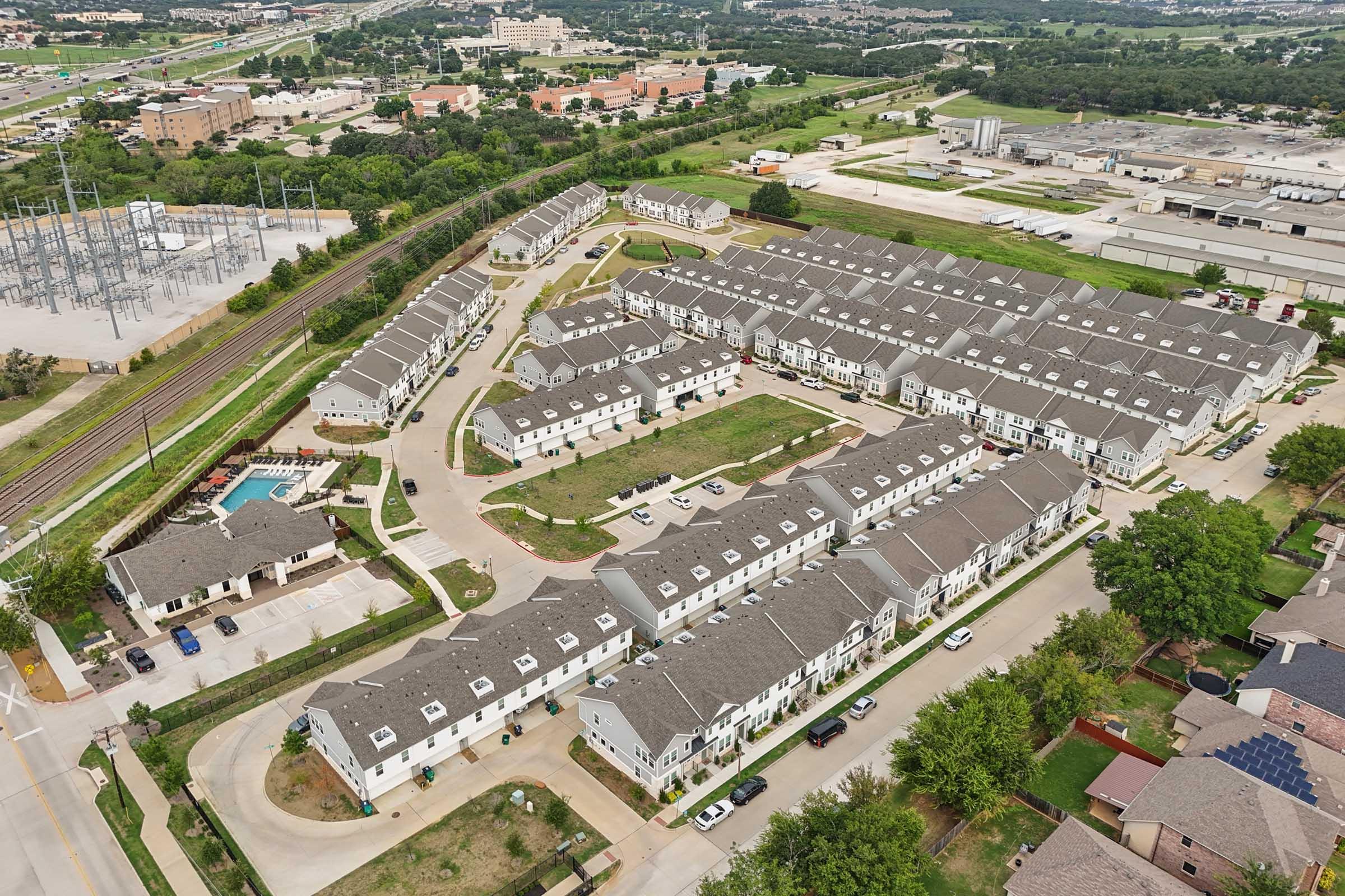 Aerial view of a residential complex featuring multiple buildings arranged in a circular layout, with green spaces, a swimming pool, and parking areas. Surrounding the complex are roads, trees, and other structures, along with industrial buildings visible in the background.