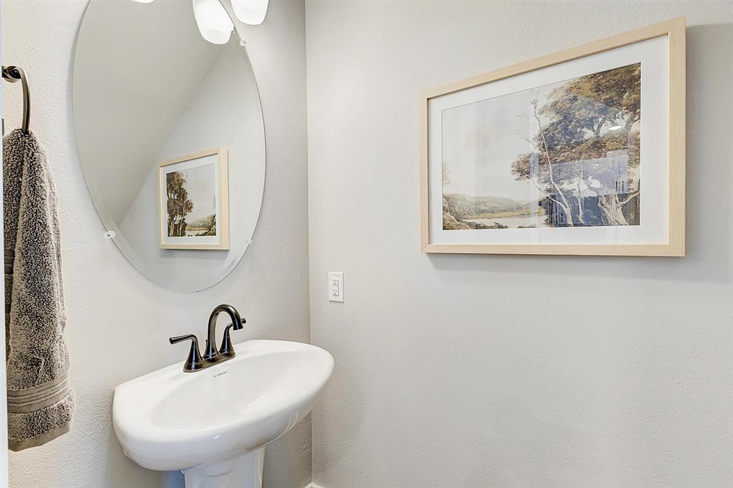 A small bathroom featuring a white sink, a round mirror above it, and a framed landscape artwork on the wall. A gray towel hangs on a hook beside the sink, with light-colored walls creating a clean and minimalist atmosphere.