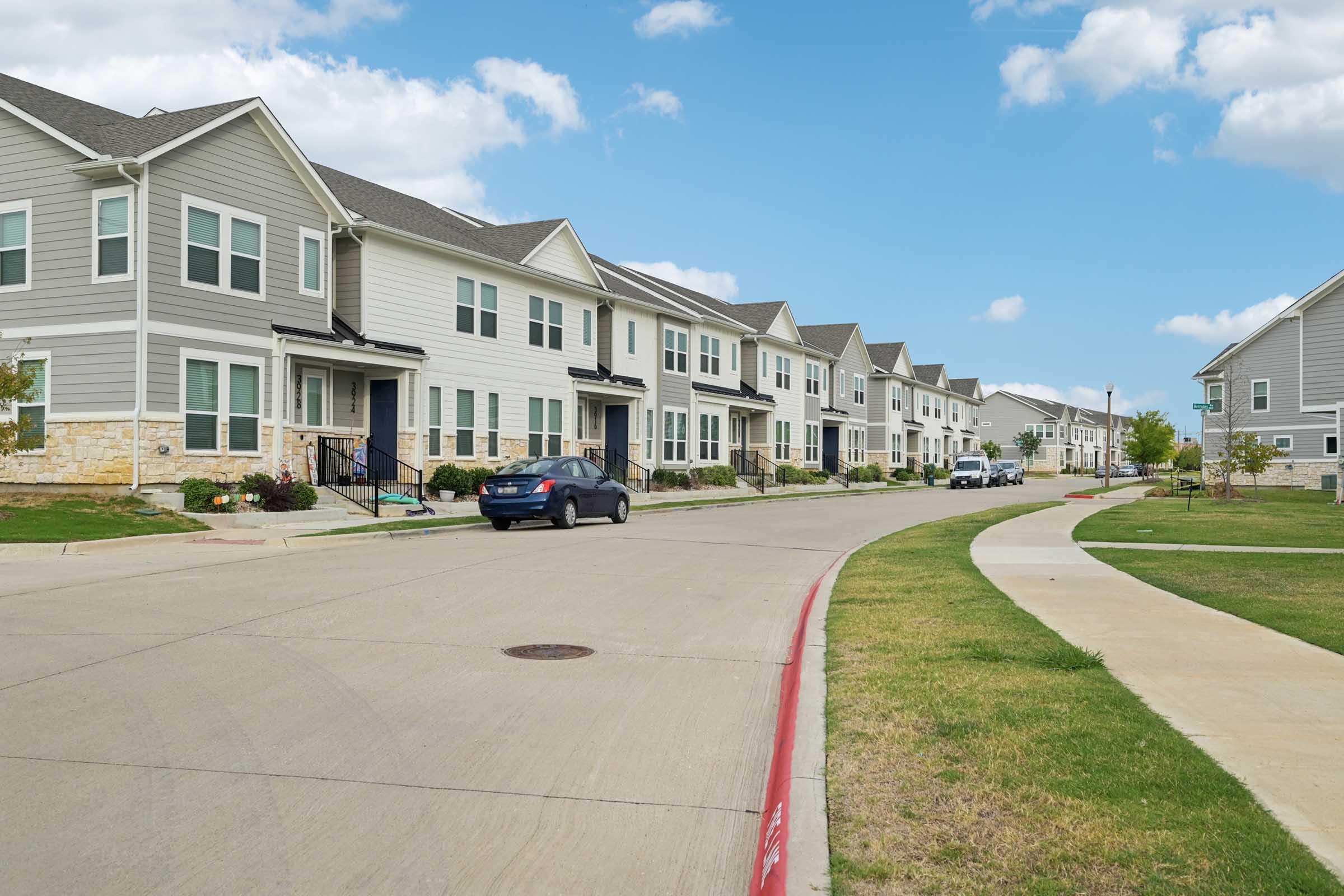 A residential street lined with modern, two-story gray and white townhouses. The road is paved and features a grassy area alongside the curb. Blue skies with a few white clouds are visible in the background, creating a pleasant suburban atmosphere.