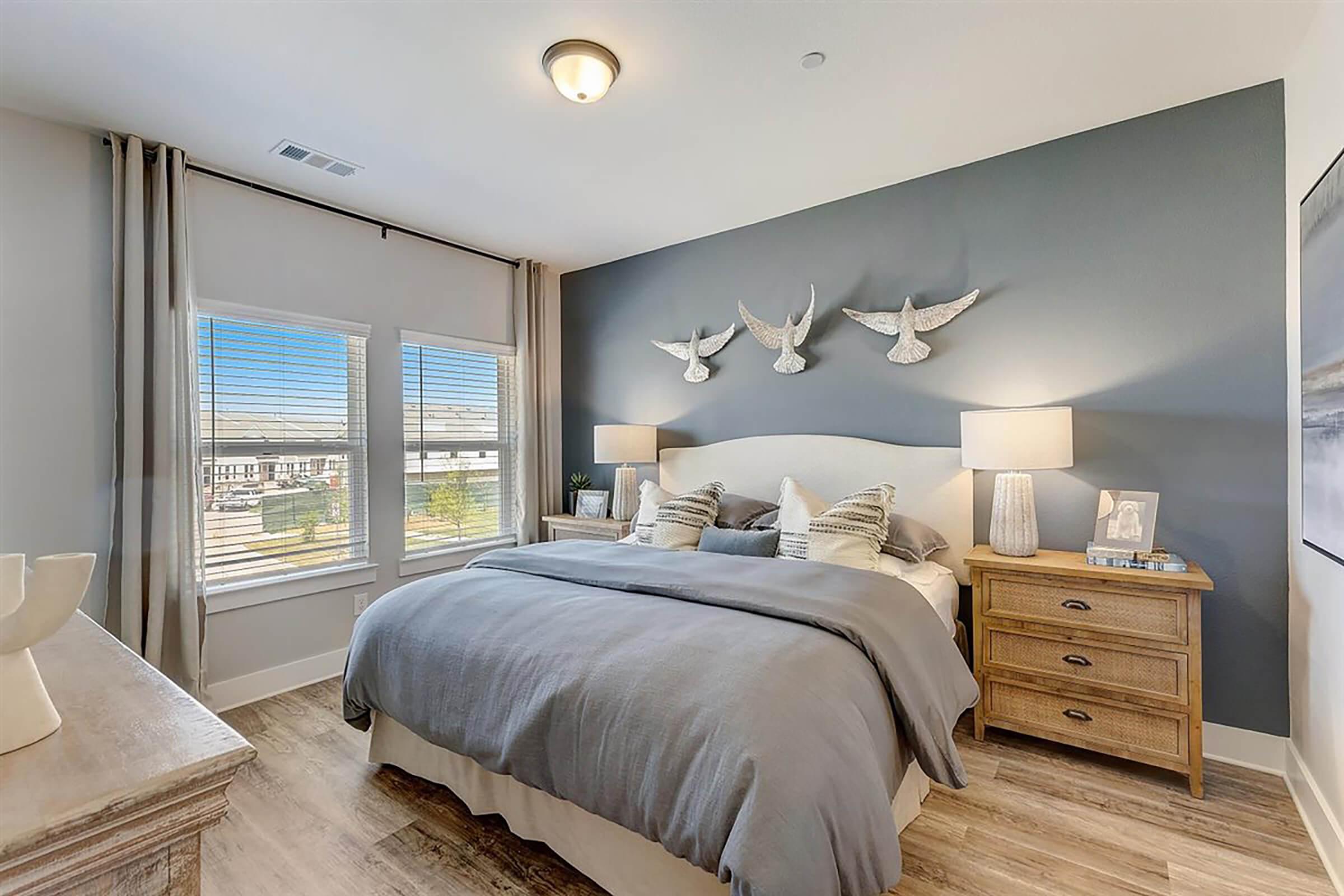 A cozy bedroom featuring a large bed with gray bedding, decorative pillows, and two bedside lamps on wooden nightstands. The walls are painted a soft gray, adorned with decorative birds, and there are large windows letting in natural light, revealing a view of the outdoors.