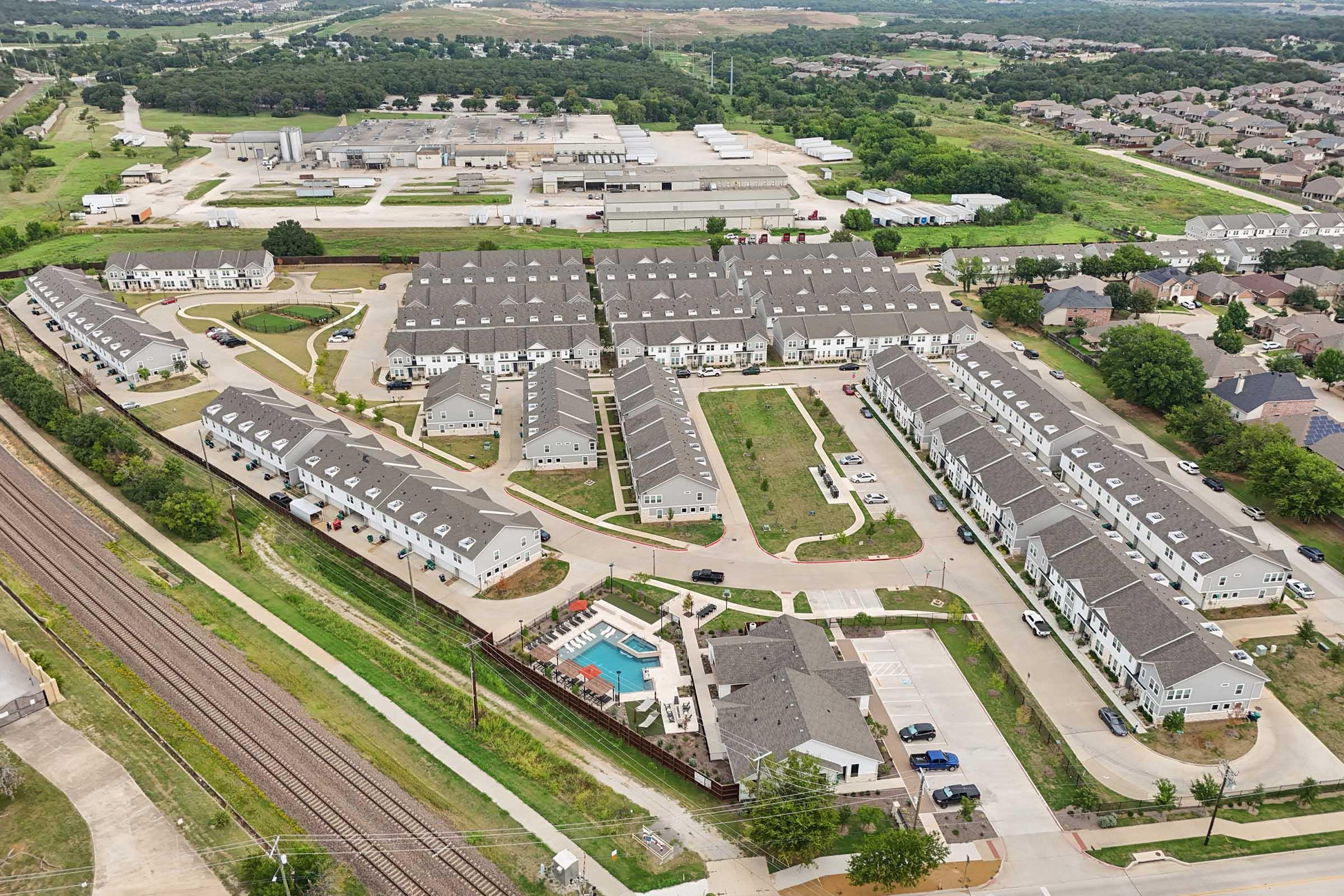 Aerial view of a residential complex featuring multiple buildings with gray roofs arranged in a circular layout. A swimming pool is visible in the center of the complex. Surrounding areas include green spaces, roads, and a railway track, with additional industrial buildings in the distance.