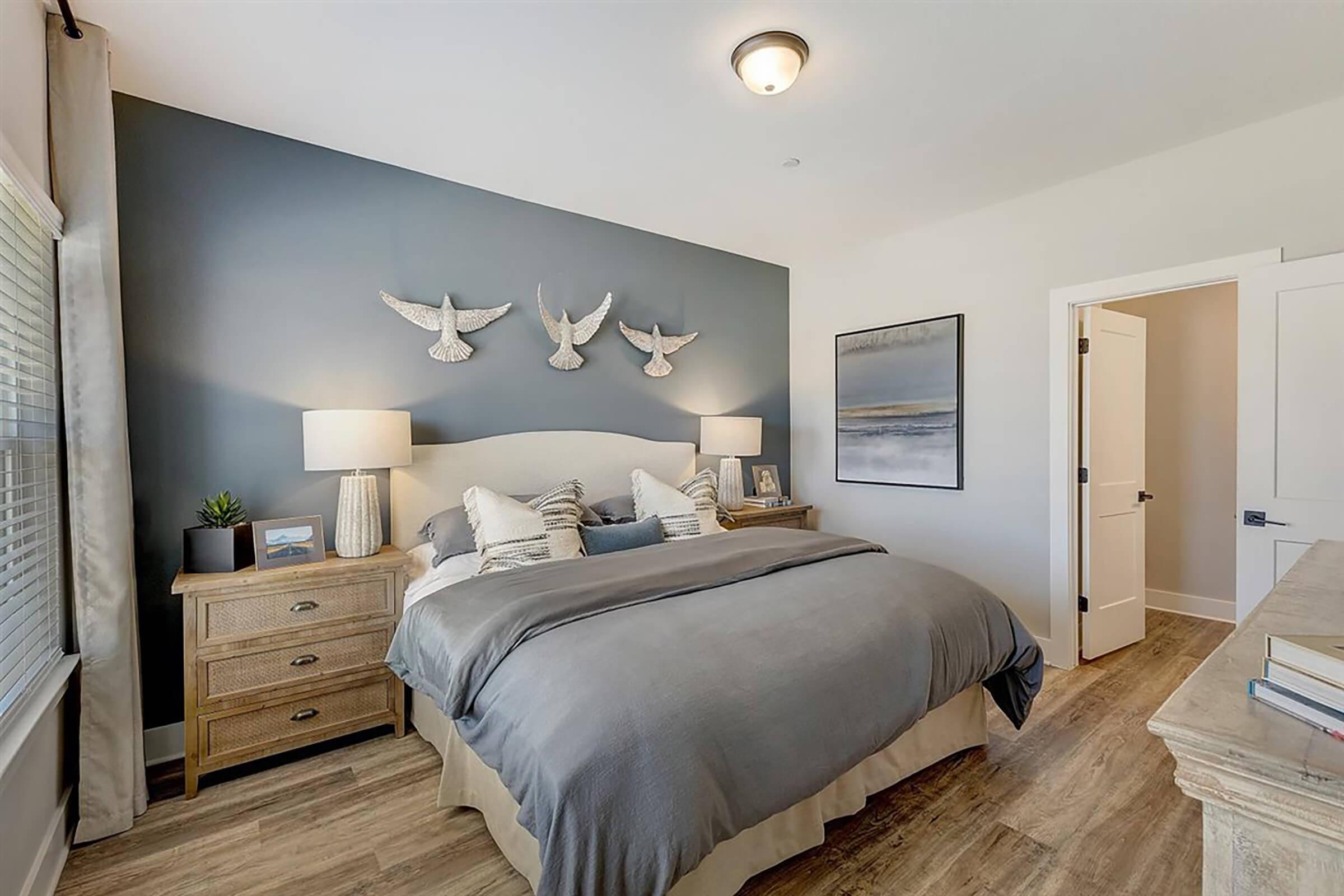 A cozy bedroom featuring a blue accent wall adorned with decorative birds. The bed is dressed in gray linens with plush pillows, flanked by two stylish bedside lamps on wooden nightstands. A piece of artwork hangs above a dresser, while a doorway leads to another room, enhancing the inviting atmosphere.