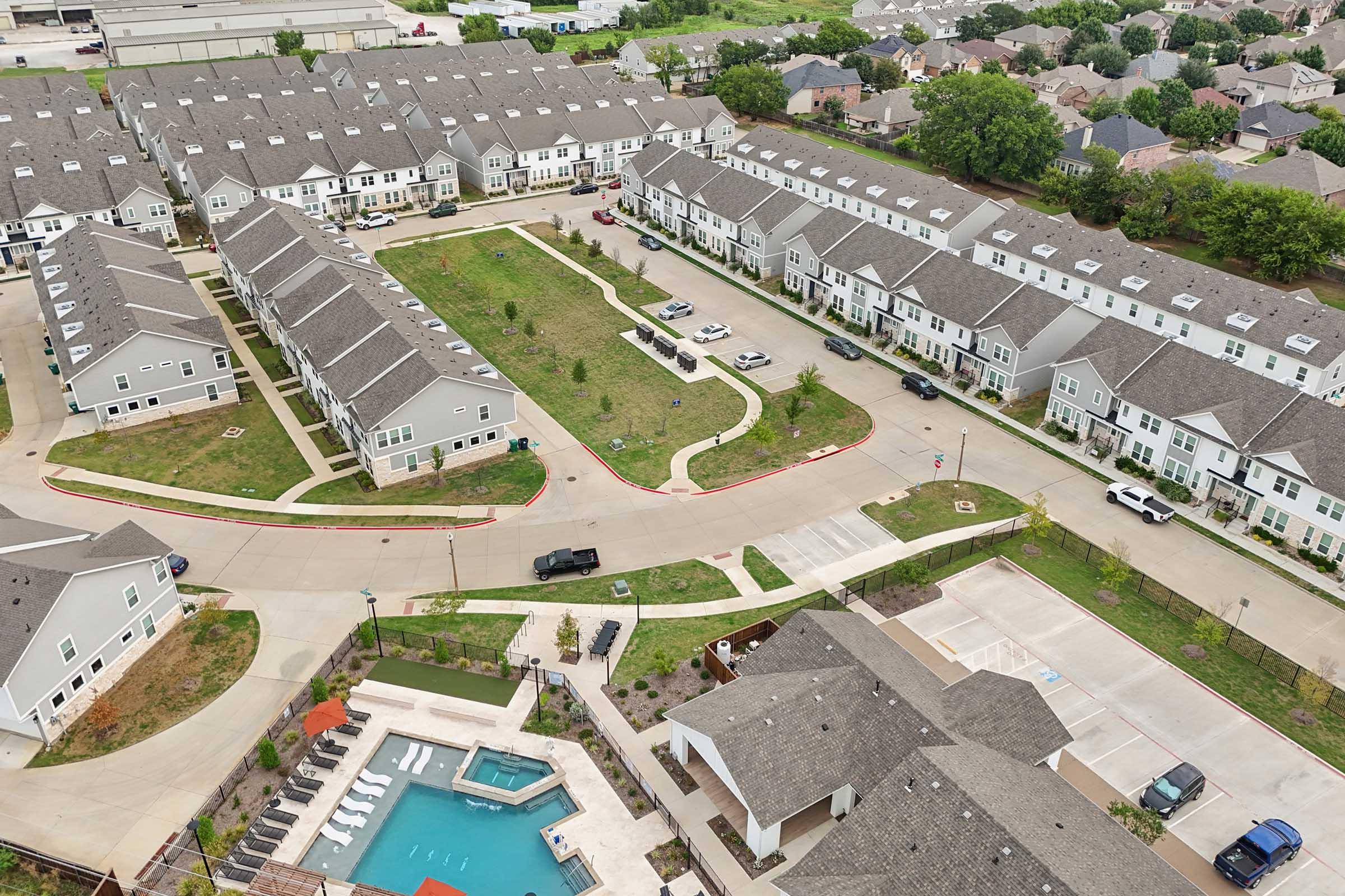Aerial view of a residential area featuring several two-story apartment buildings arranged around a central green space. A swimming pool is visible in the foreground, along with parking spaces and vehicles along the road. The neighborhood is surrounded by greenery and other homes in the background.