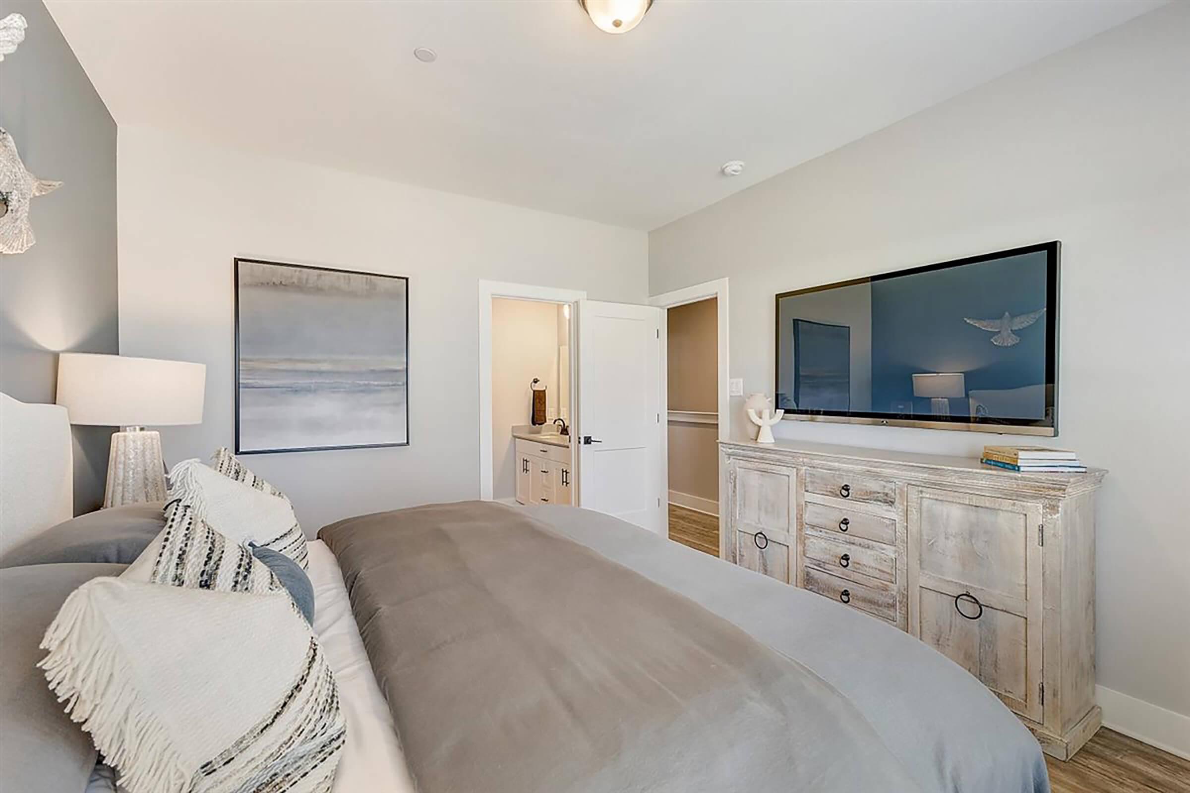 A modern bedroom featuring a cozy bed with decorative pillows, a textured gray blanket, and a light-colored accent wall. There is a large piece of artwork on the wall, a TV mounted above a wooden dresser, and a doorway leading to a bathroom. Soft lighting creates a warm atmosphere.