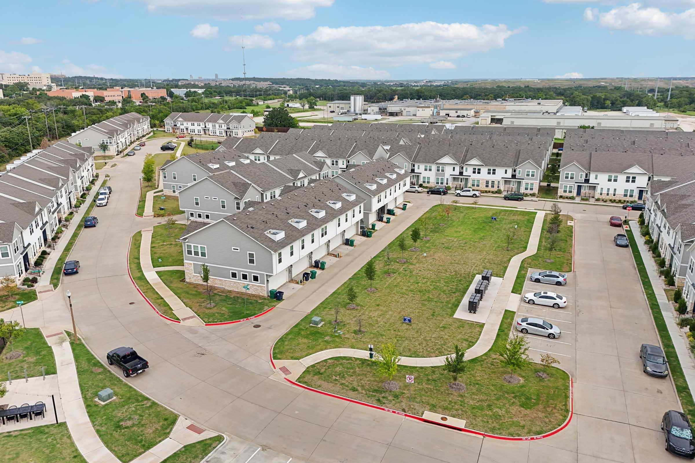 Aerial view of a residential neighborhood featuring two-story townhouses with gray roofs, arranged in a circular layout. Well-maintained green spaces and driveways are visible, alongside parked cars. The surrounding area includes additional buildings and open land, with a clear blue sky above.