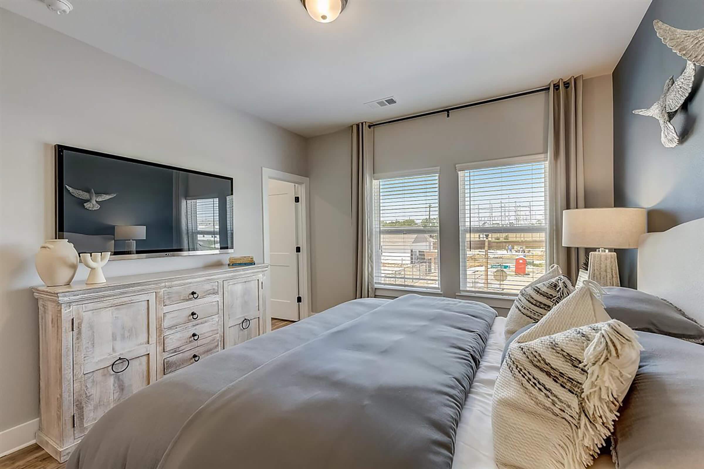 A modern bedroom featuring a neatly made bed with gray bedding, decorative pillows, and a stylish headboard. A wooden dresser with decorative items is positioned against the wall, and a flat-screen TV hangs above it. Large windows with curtain panels provide natural light and a view of the outdoors.
