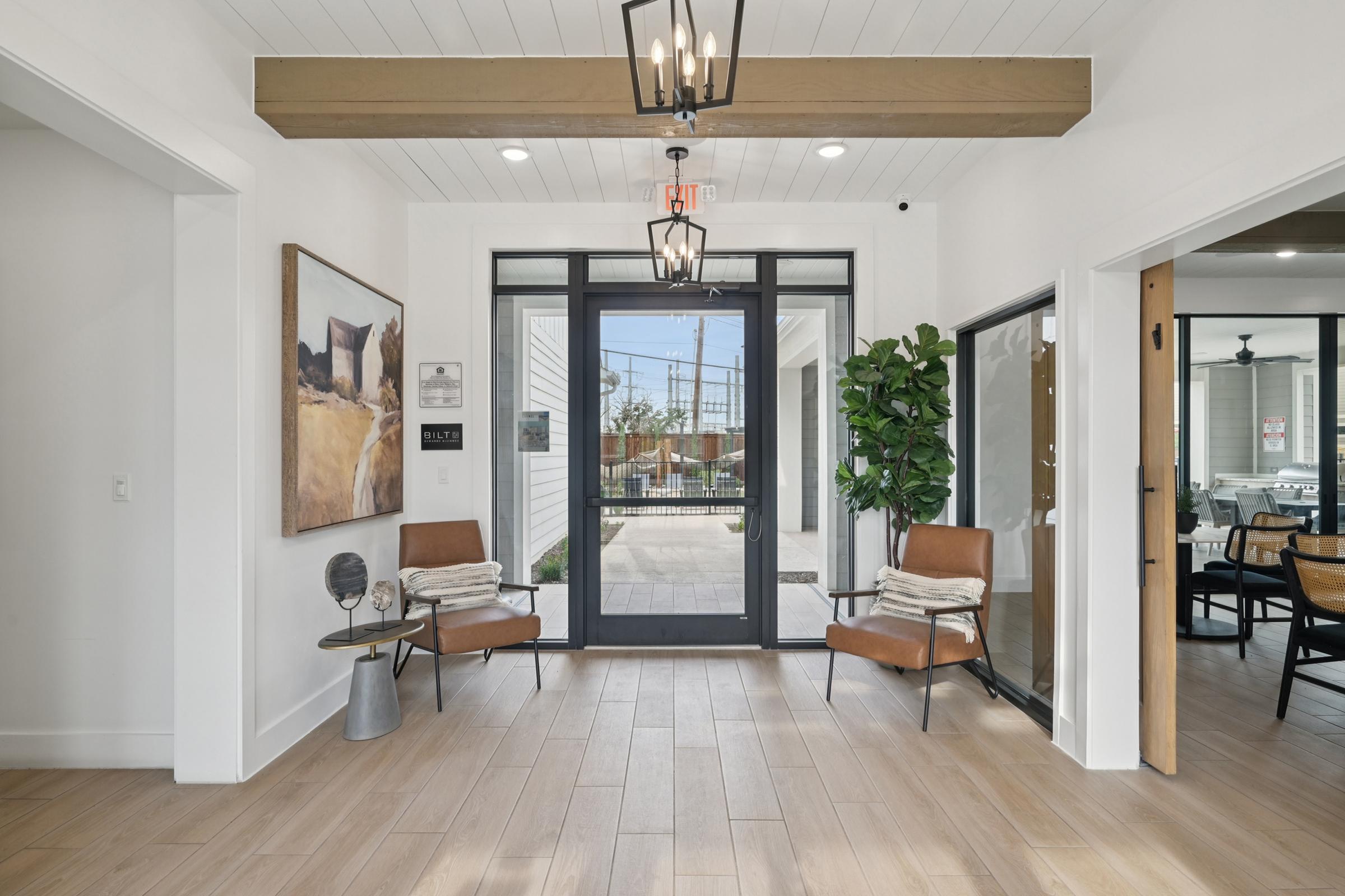 Bright and modern entryway featuring a large glass door with black frame, two brown chairs with throw blankets, a decorative plant, and a wall art piece. Natural light floods the space, highlighting the light wood flooring and the minimalist design.