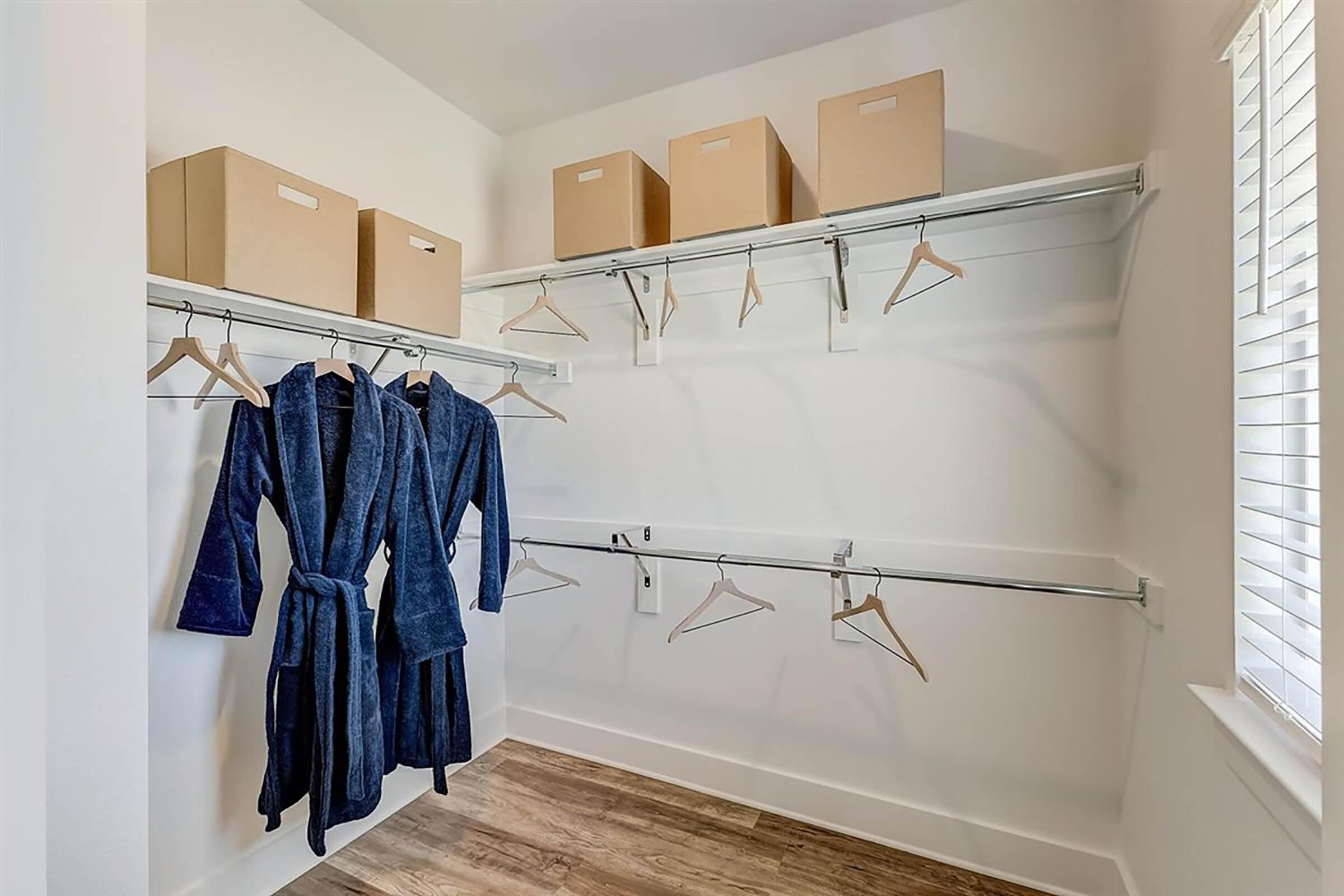A minimalist walk-in closet featuring two blue bathrobes hanging on a rod, several empty hangers, and five cardboard boxes on shelves. The walls are painted white, and there is natural light coming from a window with blinds. The floor is wooden, creating a clean and organized appearance.