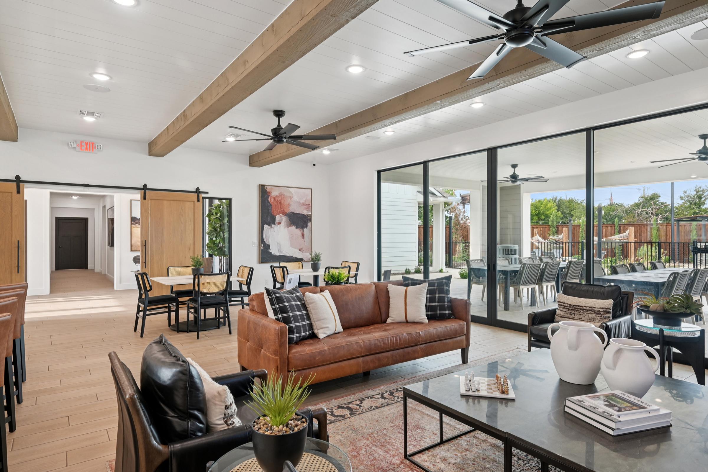 A modern interior living space featuring a leather sofa, black chairs, and a coffee table. Large windows provide natural light, showing an outdoor area. The room has wooden beams, ceiling fans, and a stylish decor with plants and artwork, creating a welcoming atmosphere for social gatherings.