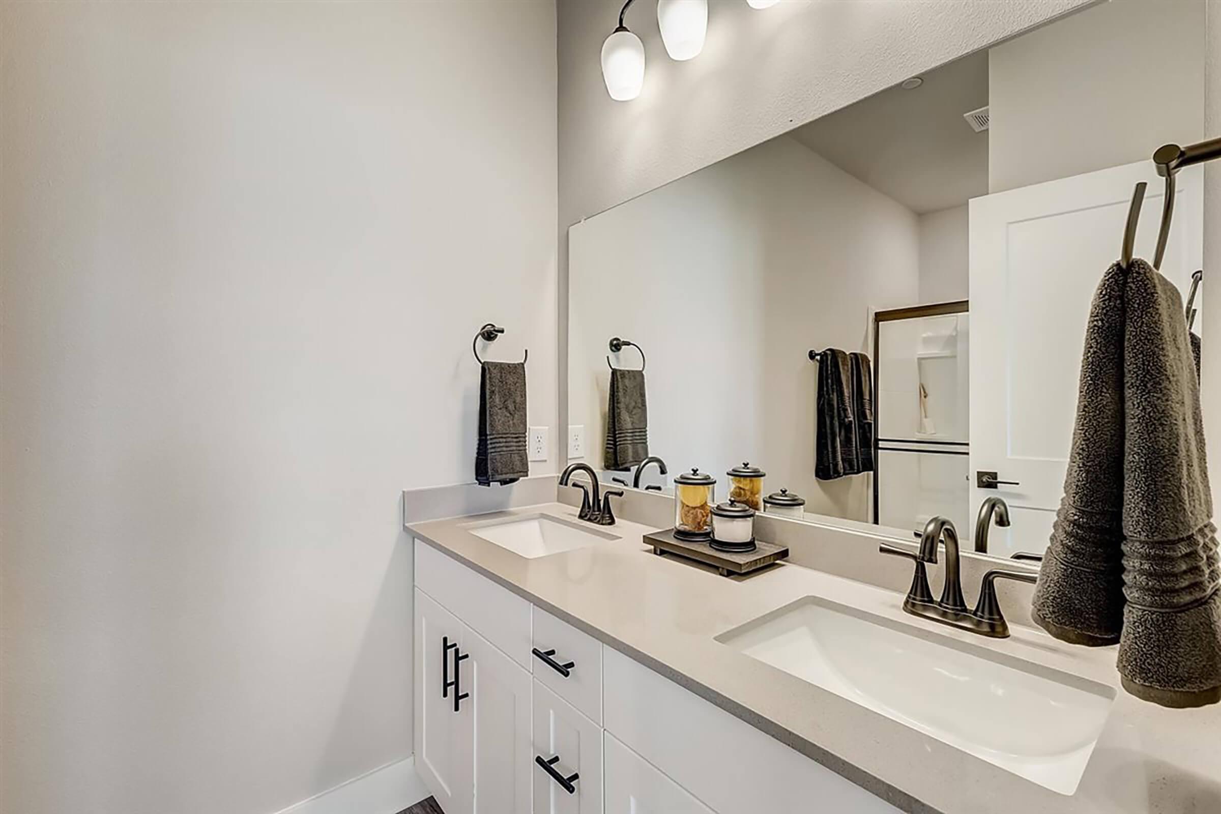 A modern bathroom featuring a double vanity with white countertops, black faucets, and two sinks. The wall is painted in a light color, and there is a large mirror above the vanity. Two towels hang on towel bars, and decorative items are arranged on the countertop. Bright lighting is provided by three pendant lights.