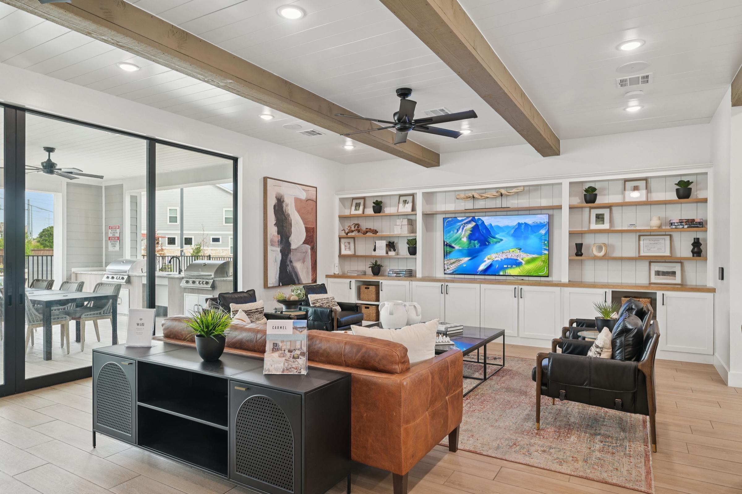 Bright and modern living room featuring a comfortable brown leather sofa, a stylish black accent chair, and a large wall-mounted TV displaying a scenic mountain landscape. Open shelves with decorative items and books, along with large windows providing natural light. A ceiling fan and light wood beams add warmth to the space.