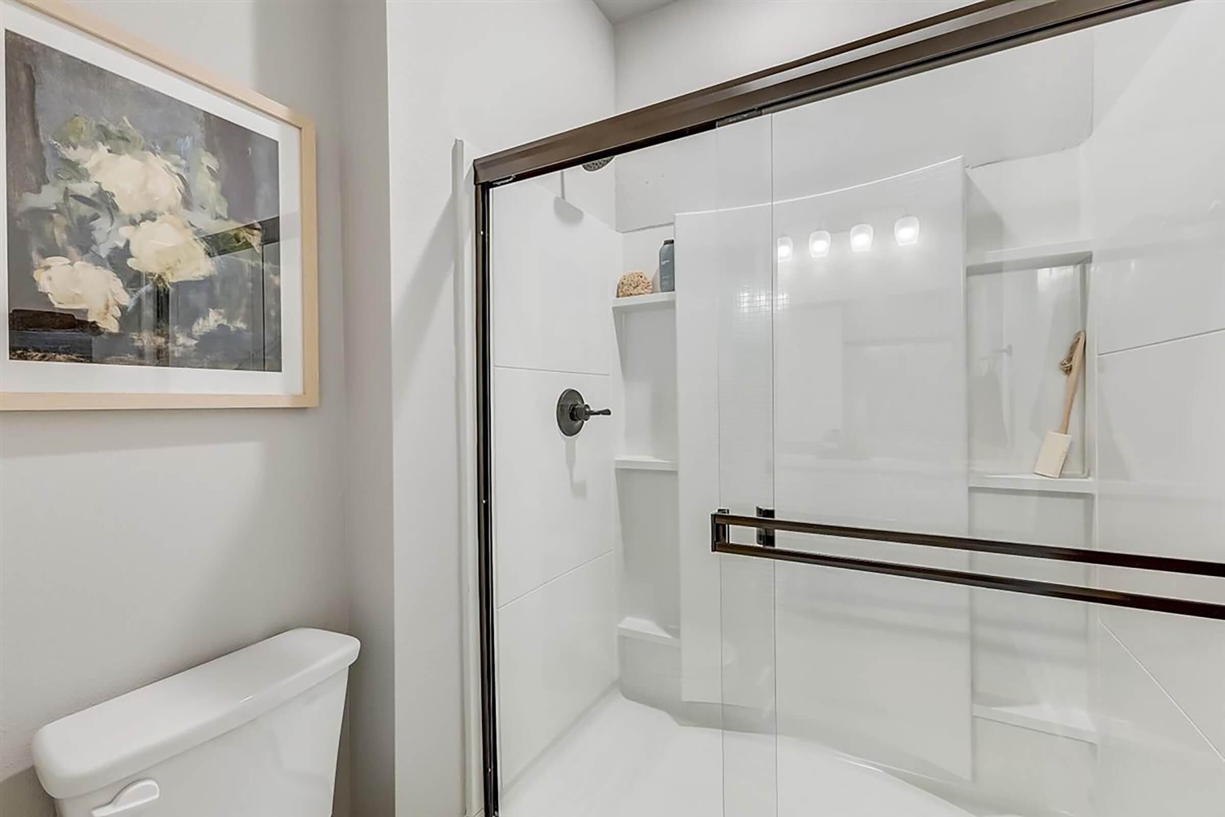 A modern bathroom featuring a glass shower enclosure, a light-colored wall, and a framed painting of flowers. There's a toilet to the left and built-in shelves in the shower area, with a few decorative items visible. The overall design is clean and contemporary.
