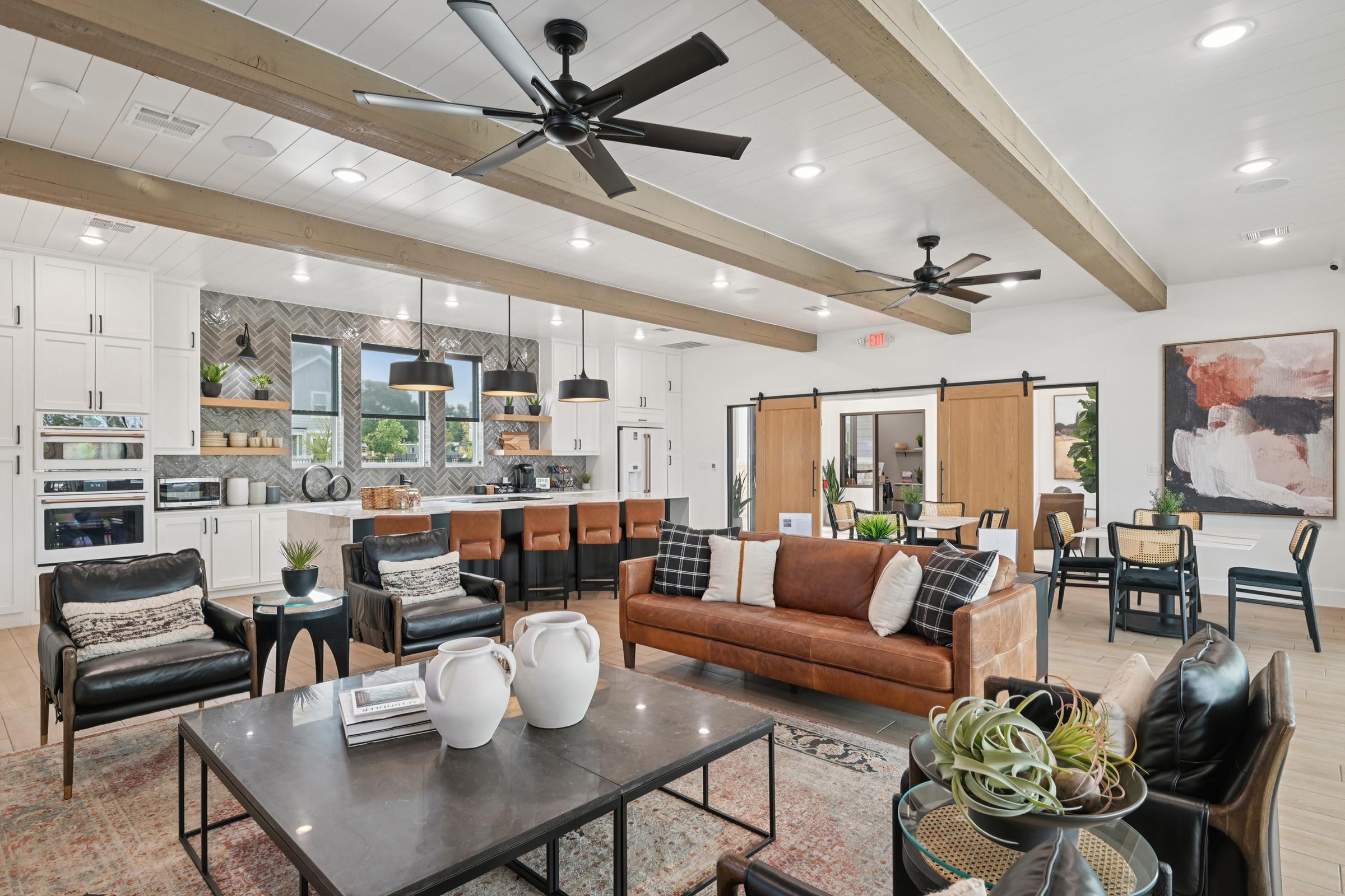 A modern living room with an open layout features a brown leather sofa, black chairs, and a coffee table. A dining area with a large table is visible in the background, along with a stylish kitchen. The space has white cabinets, wooden beams, and decorative plants, creating a warm and inviting atmosphere.