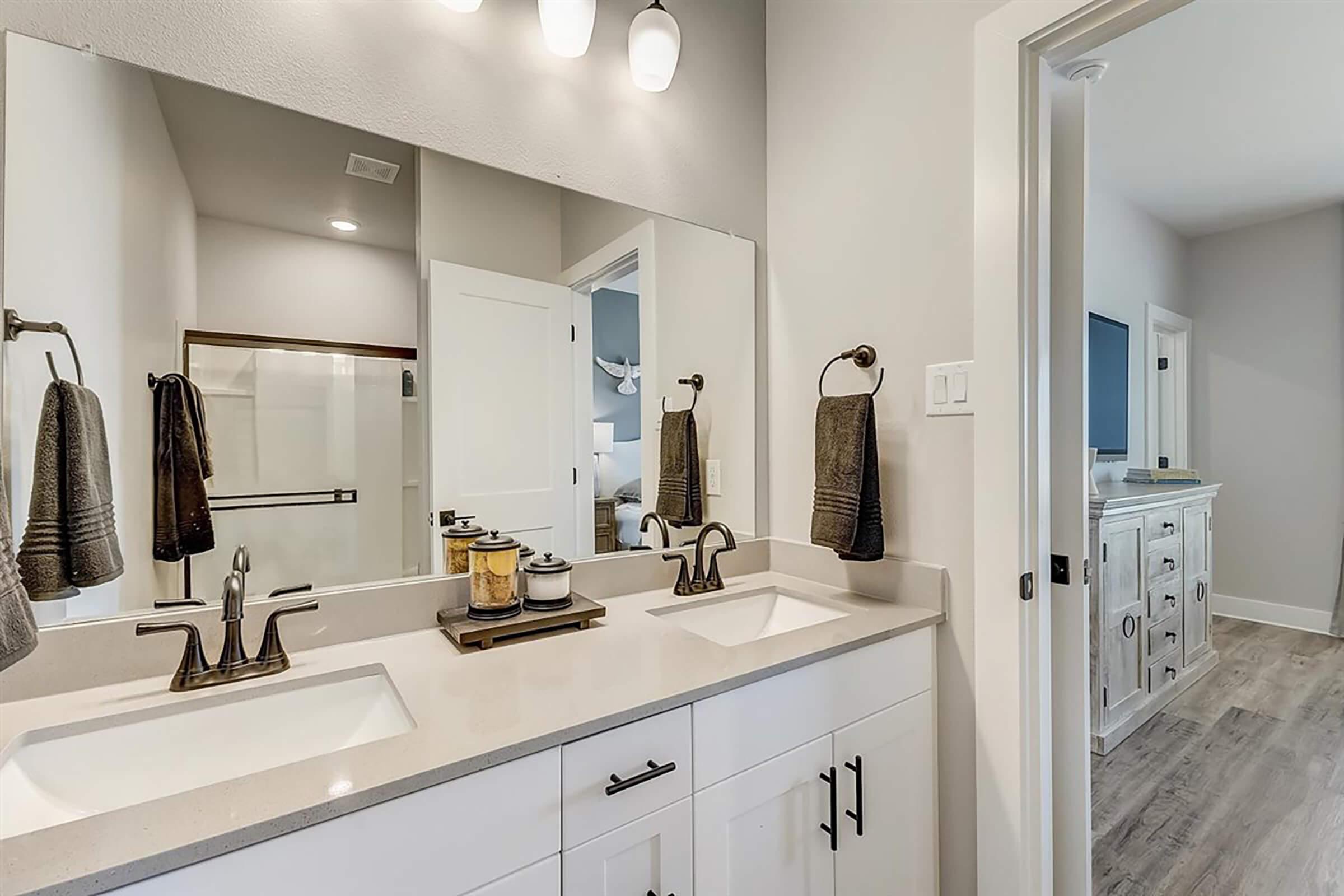 A modern bathroom featuring a double vanity with two sinks, elegant faucets, and decorative accessories. The walls are painted in soft gray, and there is a large mirror above the vanity. Towel bars with gray towels are attached to the wall, and a doorway leads to another room. Natural light brightens the space.