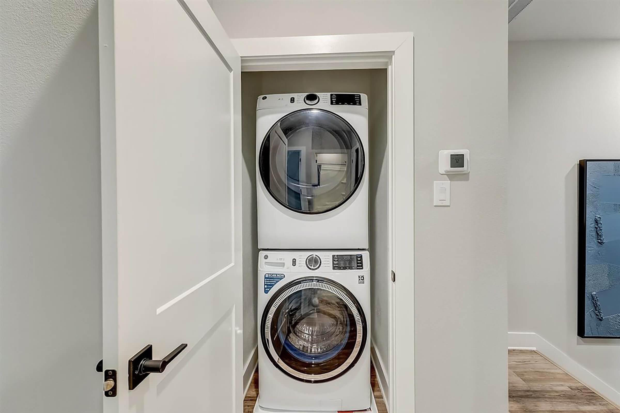 Stacked washer and dryer in a small closet space, with a white door that is partially open. The appliances are modern and sleek, featuring digital controls. The surrounding walls are painted in a light color, and there is a thermostat on the wall next to the appliances.