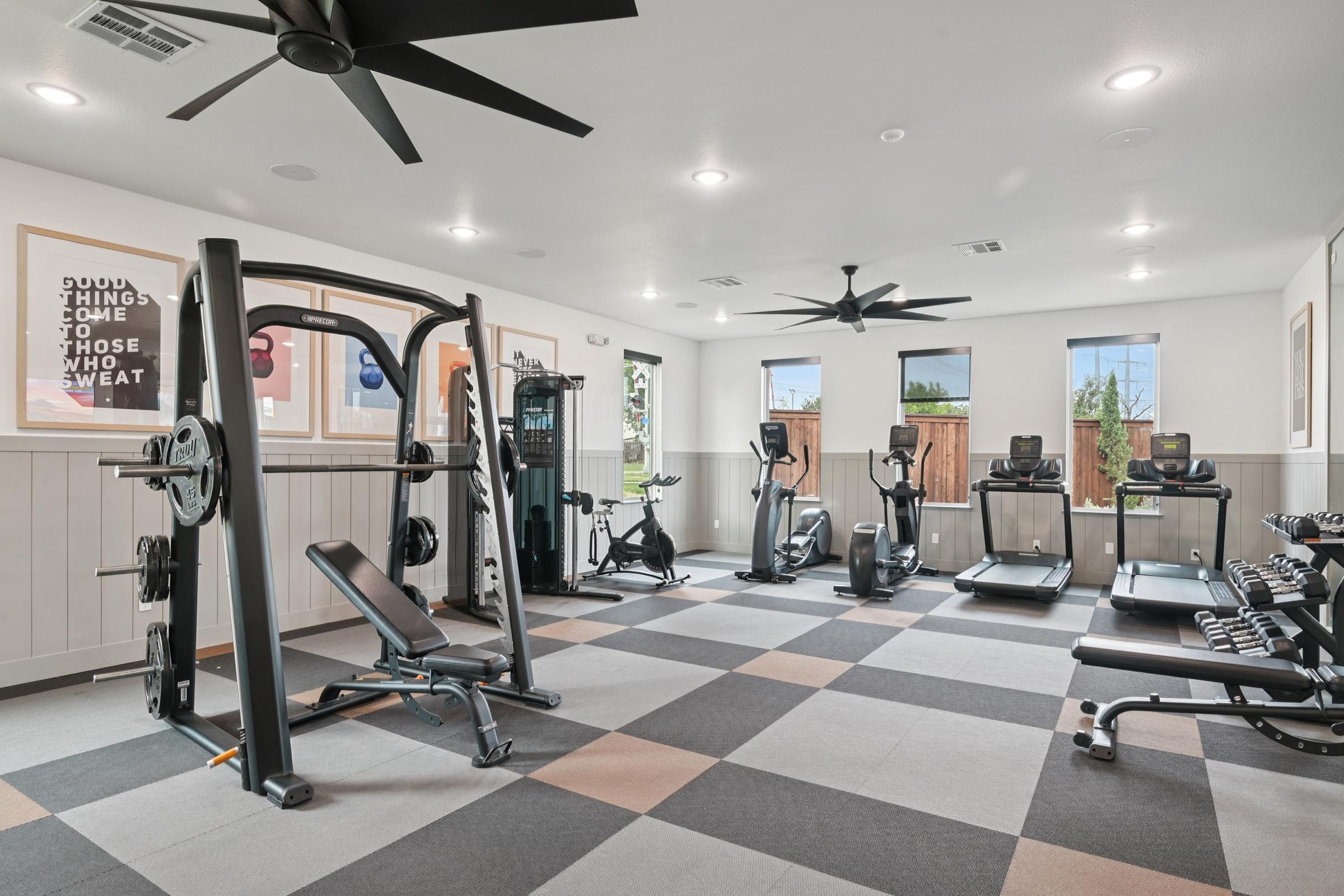 A modern gym interior featuring exercise equipment such as weights, a bench press, and cardio machines. The room has large windows allowing natural light, with a neutral color scheme and a motivational poster on the wall. The flooring is a textured pattern with gray and black tiles.