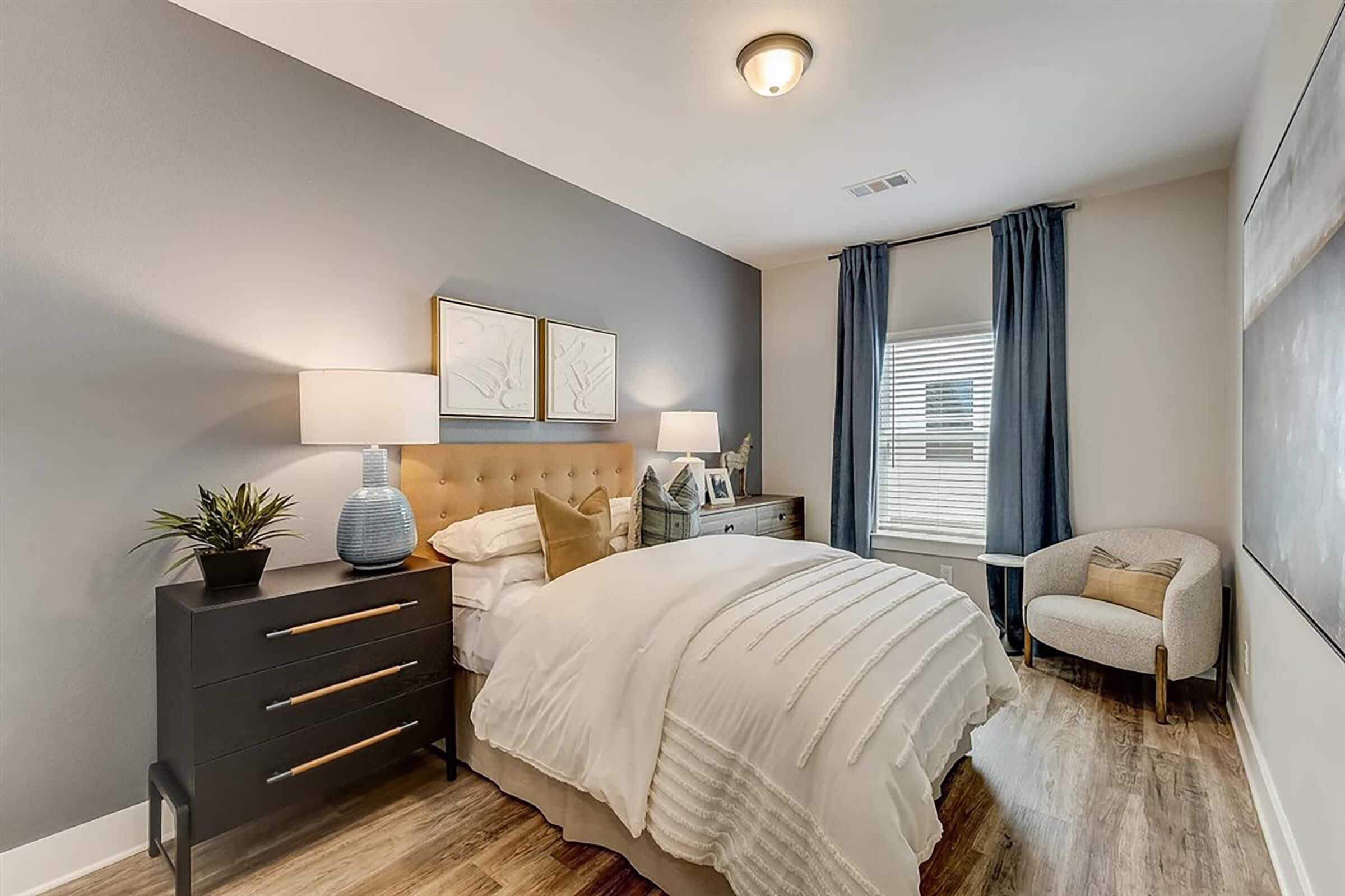 A cozy bedroom featuring a tufted beige headboard, white bedding, and two decorative pillows. There are two bedside lamps on a stylish black dresser and a potted plant on top. The room has light gray walls, a large window with blue curtains, and a comfortable armchair in the corner, enhancing the inviting atmosphere.