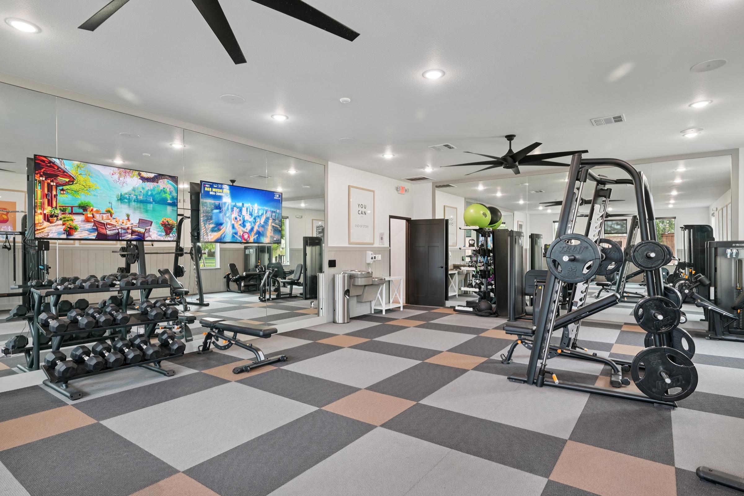 A modern gym interior featuring a workout area with dumbbells, weight machines, and exercise equipment. Two large TVs display fitness content, and the space has a sleek tile floor with a mix of colors. Large mirrors reflect the bright, well-lit environment, creating an inviting atmosphere for exercising.