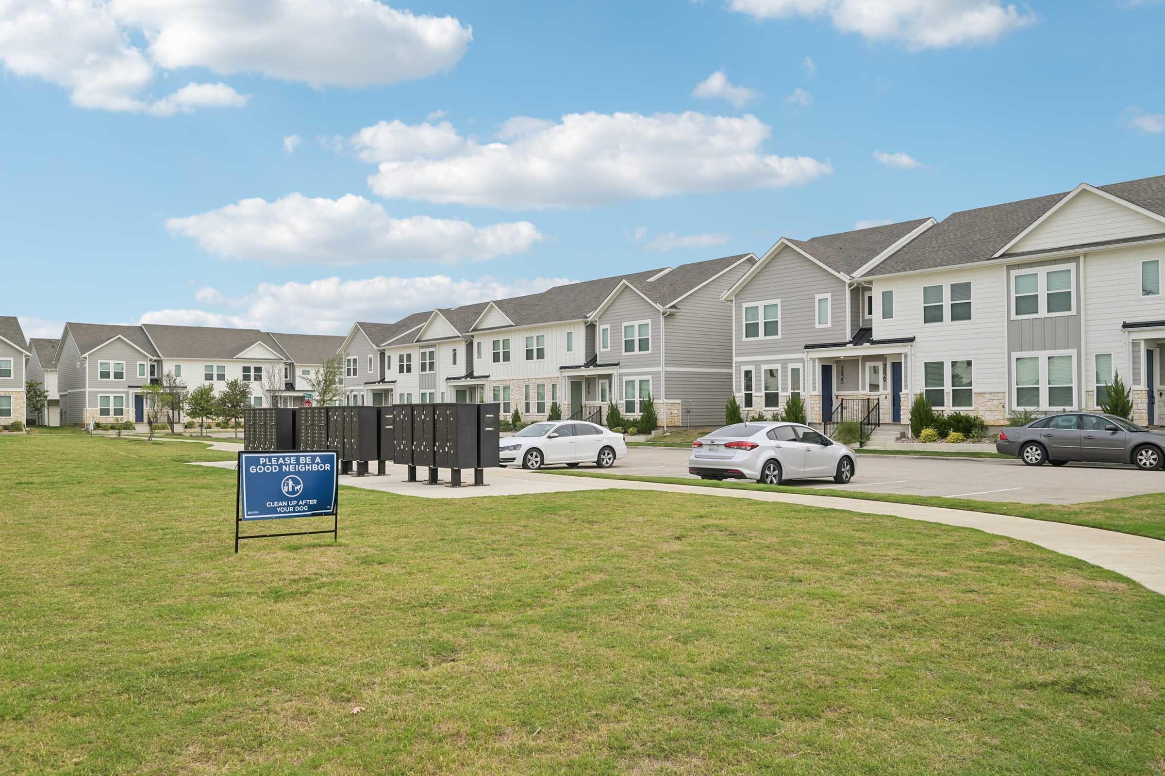 A view of a residential area with several modern townhouses and a well-maintained lawn. Parked cars line the driveway, and a sign promoting leasing options is visible in the foreground. The sky is blue with a few clouds, creating a welcoming atmosphere.