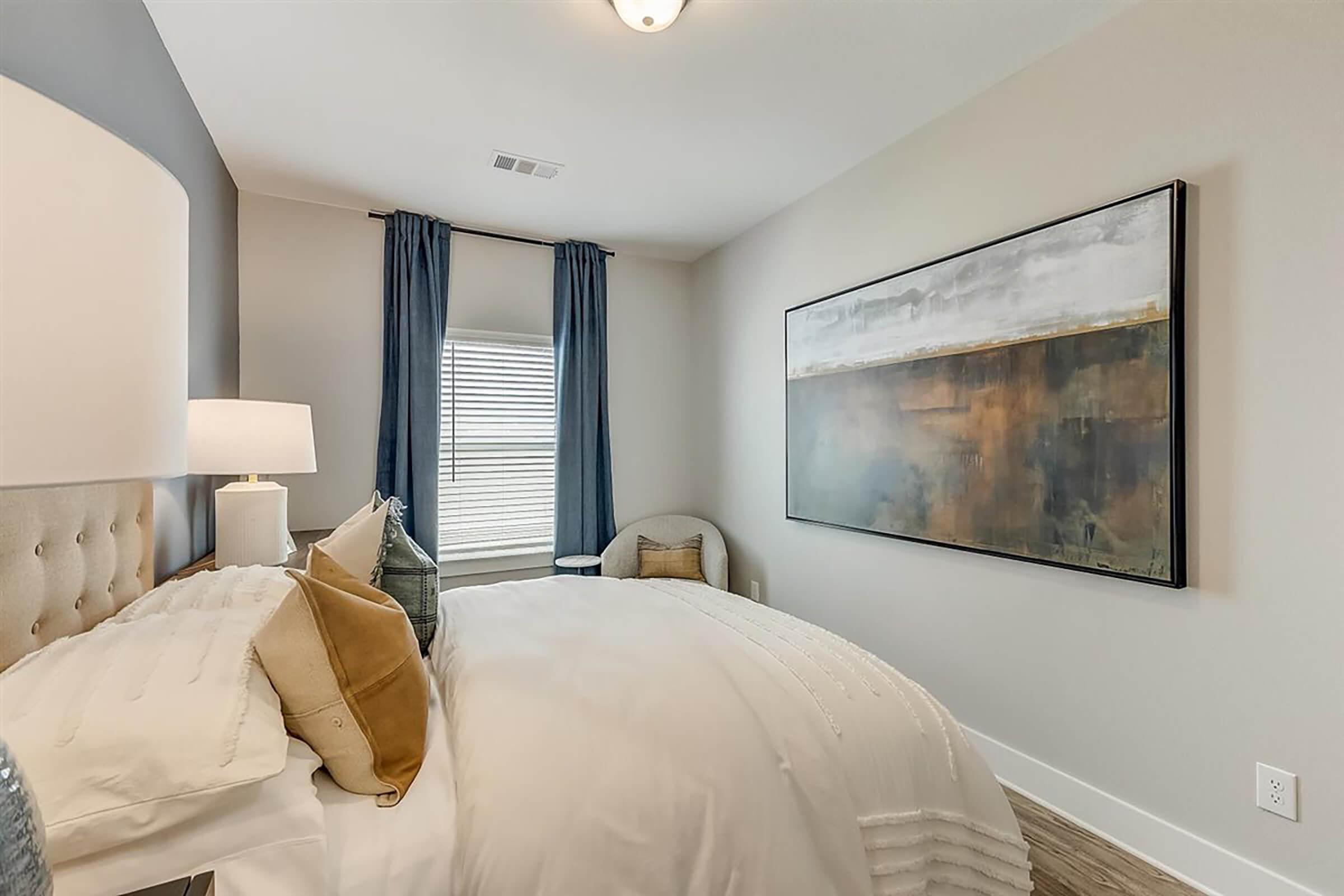 A cozy bedroom featuring a neatly made bed with white bedding and decorative pillows. There's a stylish lamp on a bedside table, blue curtains on the window, and a large abstract painting on the wall. The room has neutral tones with warm accents and hardwood flooring, creating a welcoming atmosphere.