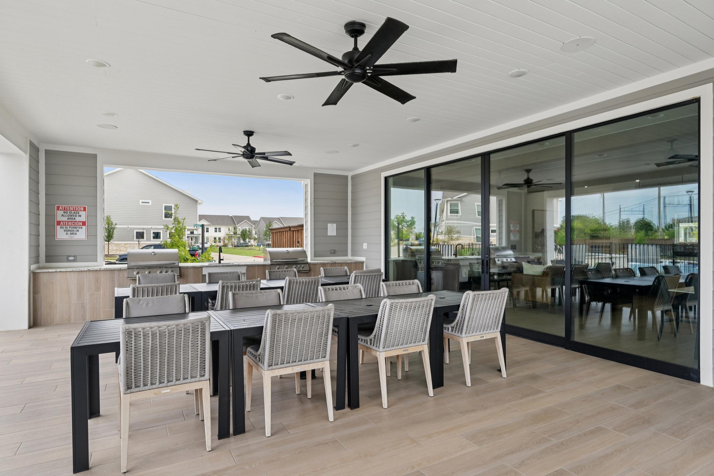 An outdoor dining area featuring a large black table surrounded by several gray chairs. The space includes a built-in grill area, ceiling fans, and large glass doors that provide a view of a grassy area and nearby buildings. The floor is tiled, and the setting is bright and welcoming.
