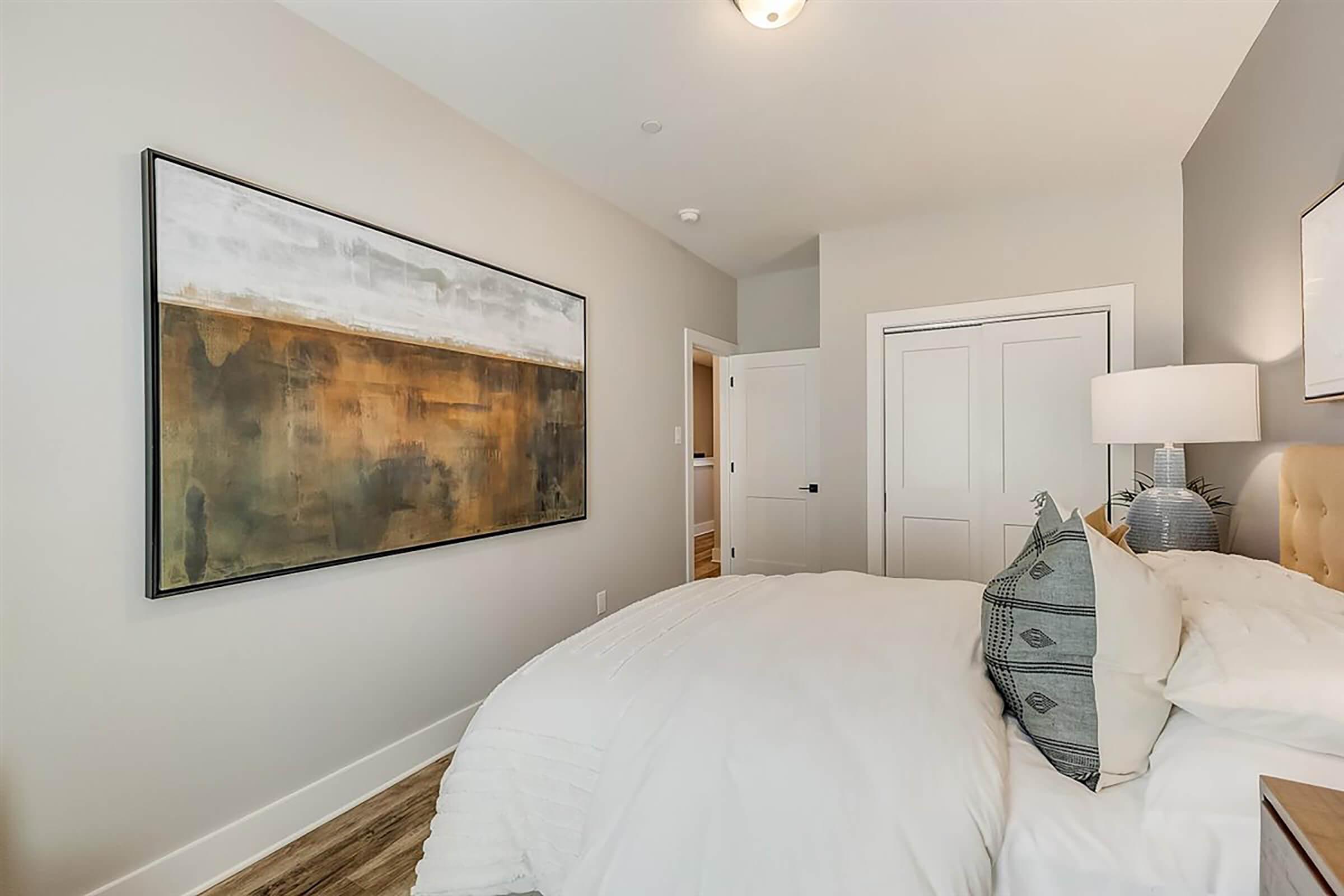 A cozy bedroom featuring a large abstract painting on the wall, a neatly made bed with white linens and decorative pillows, and a bedside lamp. The walls are painted in soft tones, and there is a door leading to another room in the background, enhancing the inviting atmosphere.