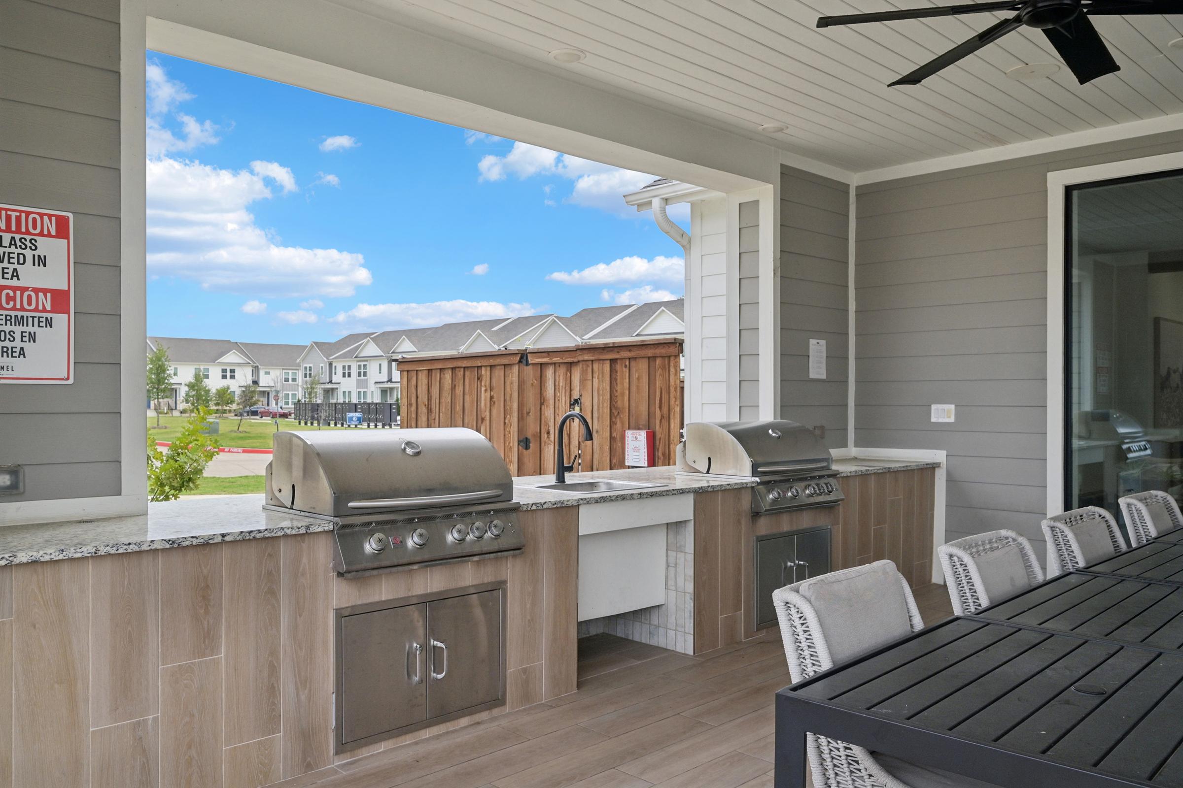 Outdoor kitchen with two stainless steel grills and a sink, set against a backdrop of green grass and blue sky. A large dining table with gray chairs is visible in the foreground, while a nearby sign indicates safety instructions. Modern residential buildings line the background.