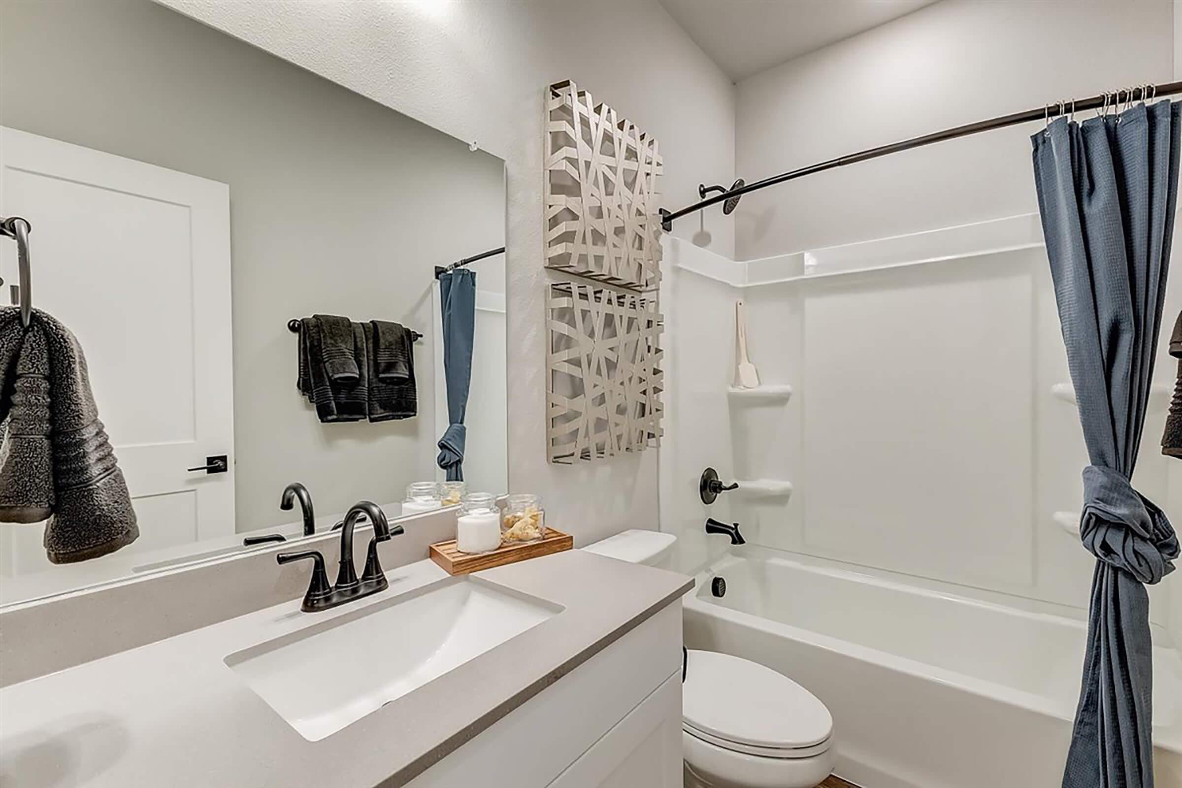 A modern bathroom featuring a white bathtub, a dark faucet, and a gray countertop. There are towels hanging on a rack, and decorative items on the countertop. The walls are light gray, and a large mirror reflects the space. A blue shower curtain adds a pop of color to the overall neutral design.
