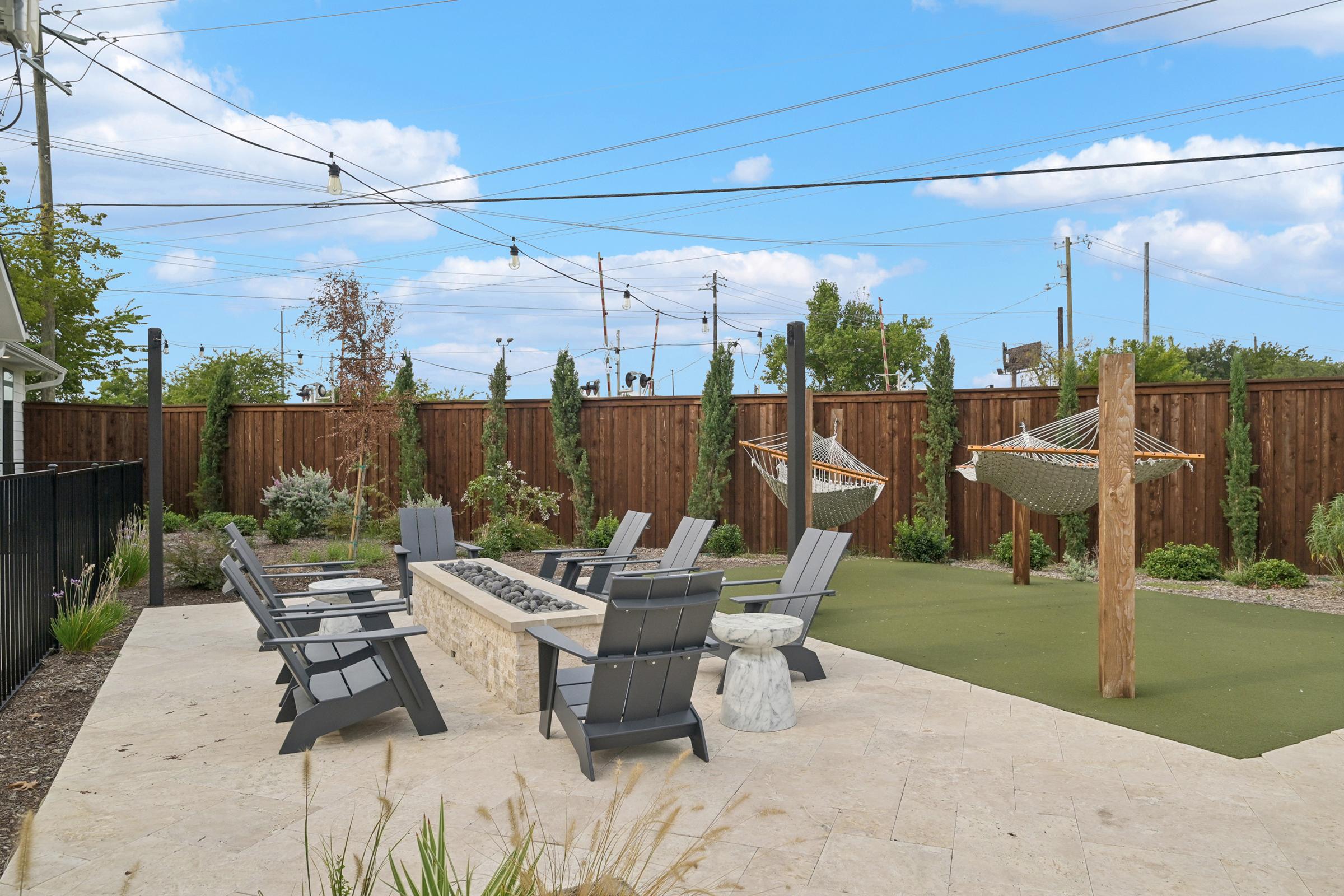 A landscaped outdoor area featuring a circular stone fire pit surrounded by gray Adirondack chairs. Two hammocks are suspended between wooden posts, and the space is bordered by a wooden fence with trees and shrubs in the background. The sky is blue with scattered clouds.