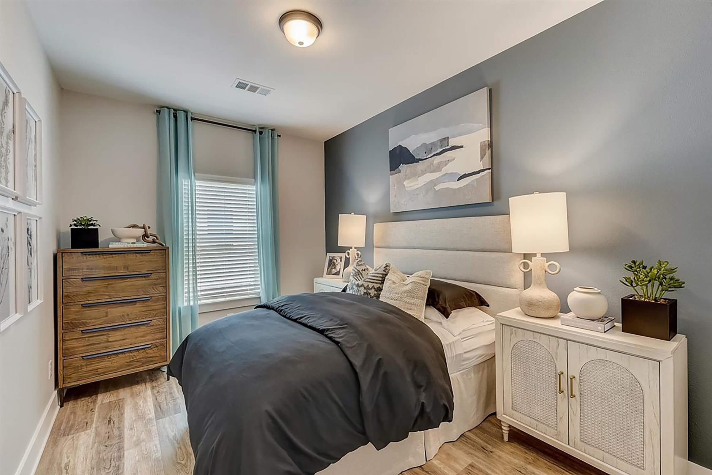A cozy bedroom featuring a light gray accent wall, a queen-sized bed with dark bedding, and decorative pillows. There are two bedside lamps on matching nightstands, a tall wooden dresser, and framed artwork on the walls. Natural light streams through a window with sheer curtains.