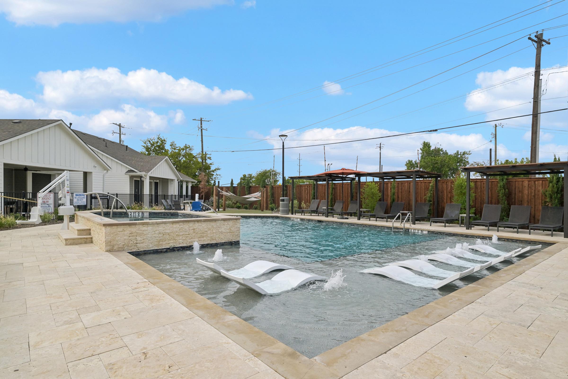 A spacious outdoor pool area featuring a large swimming pool with water jets, loungers partially submerged in the water, and a hot tub. Surrounding the pool are lounge chairs and cabanas. The sky is blue with scattered clouds, and there are residential buildings in the background.