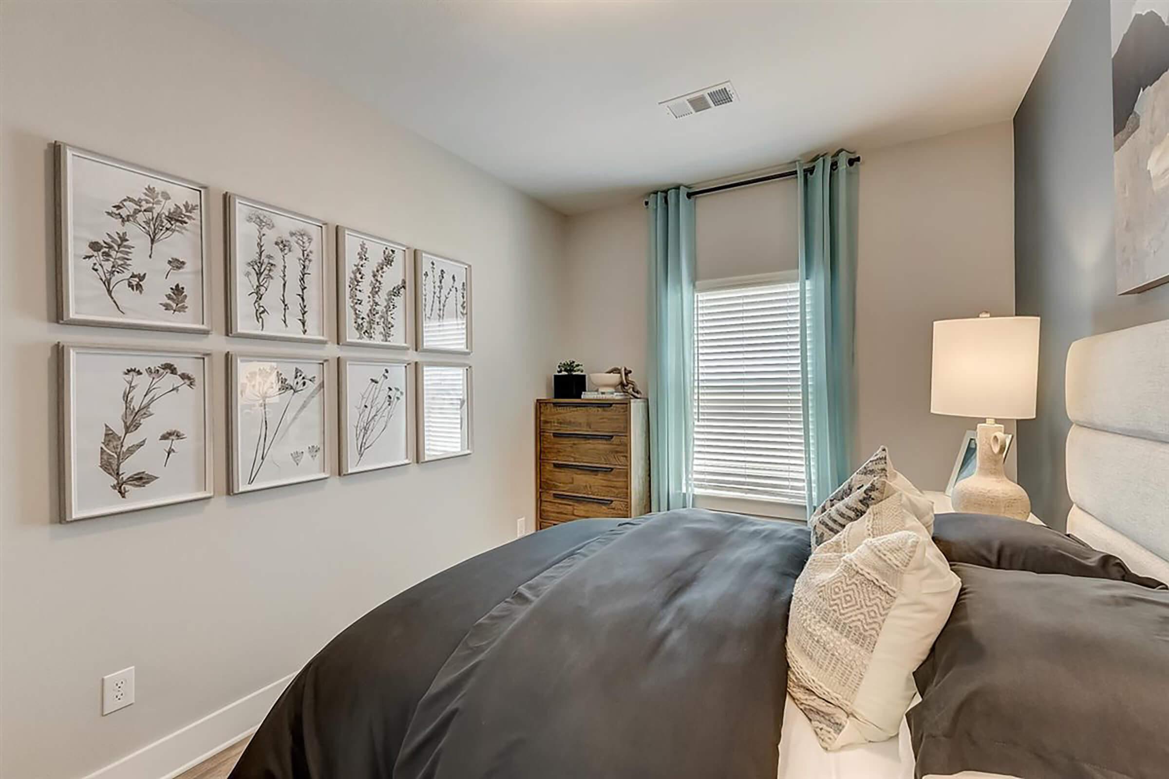 A cozy bedroom featuring a neatly made bed with dark bedding, several decorative pillows, and a soft headboard. A lamp stands on a nightstand beside the bed, and there are framed botanical prints on the wall. Light curtains frame a window, adding a serene touch to the space. A wooden dresser is visible in the corner.
