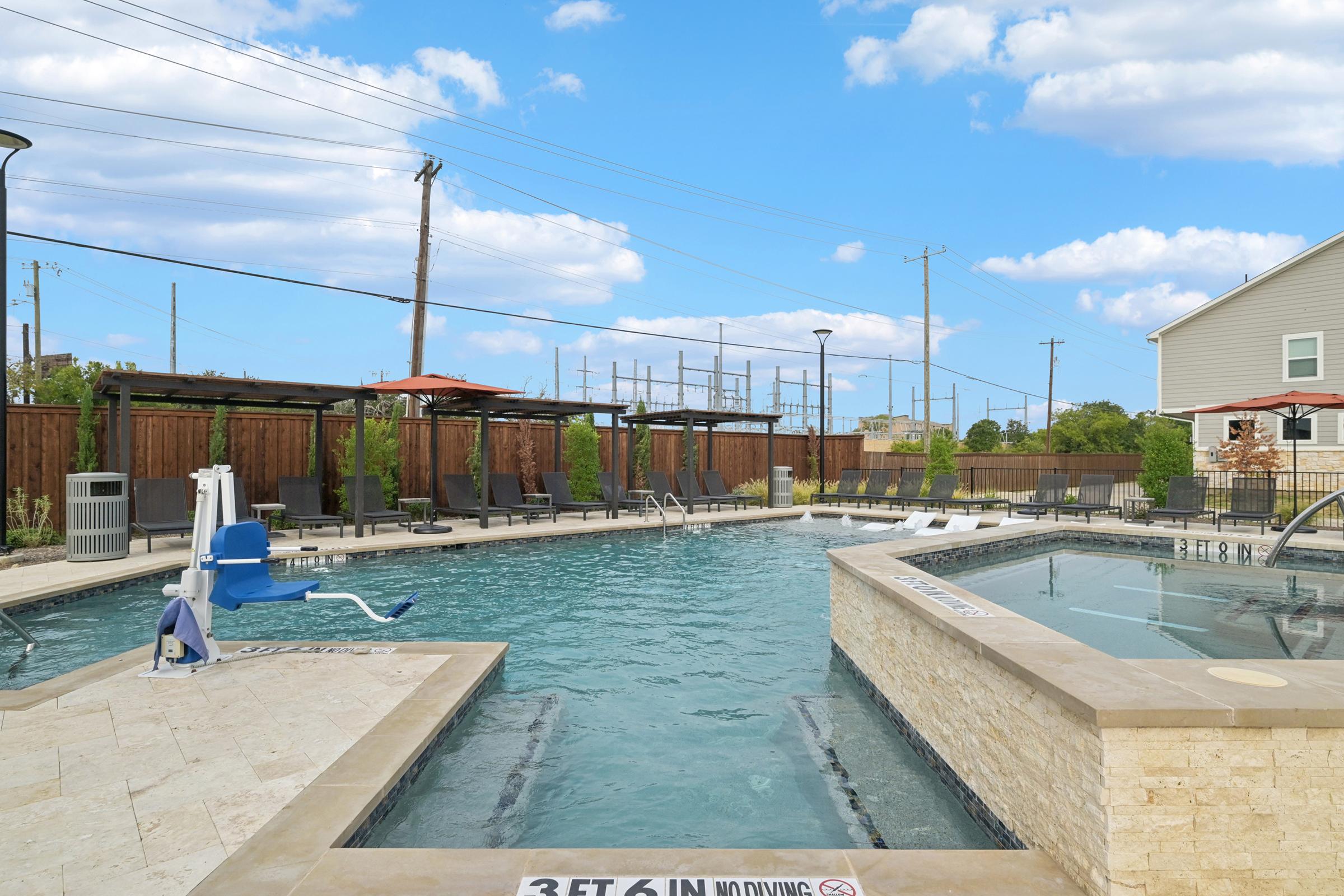 A large outdoor swimming pool featuring a hot tub, lounge chairs, and a pool lift for accessibility. The area is surrounded by wooden fencing and landscaped greenery, with a clear blue sky and scattered clouds in the background. A sign indicates the depth of the pool at 3 feet 6 inches.