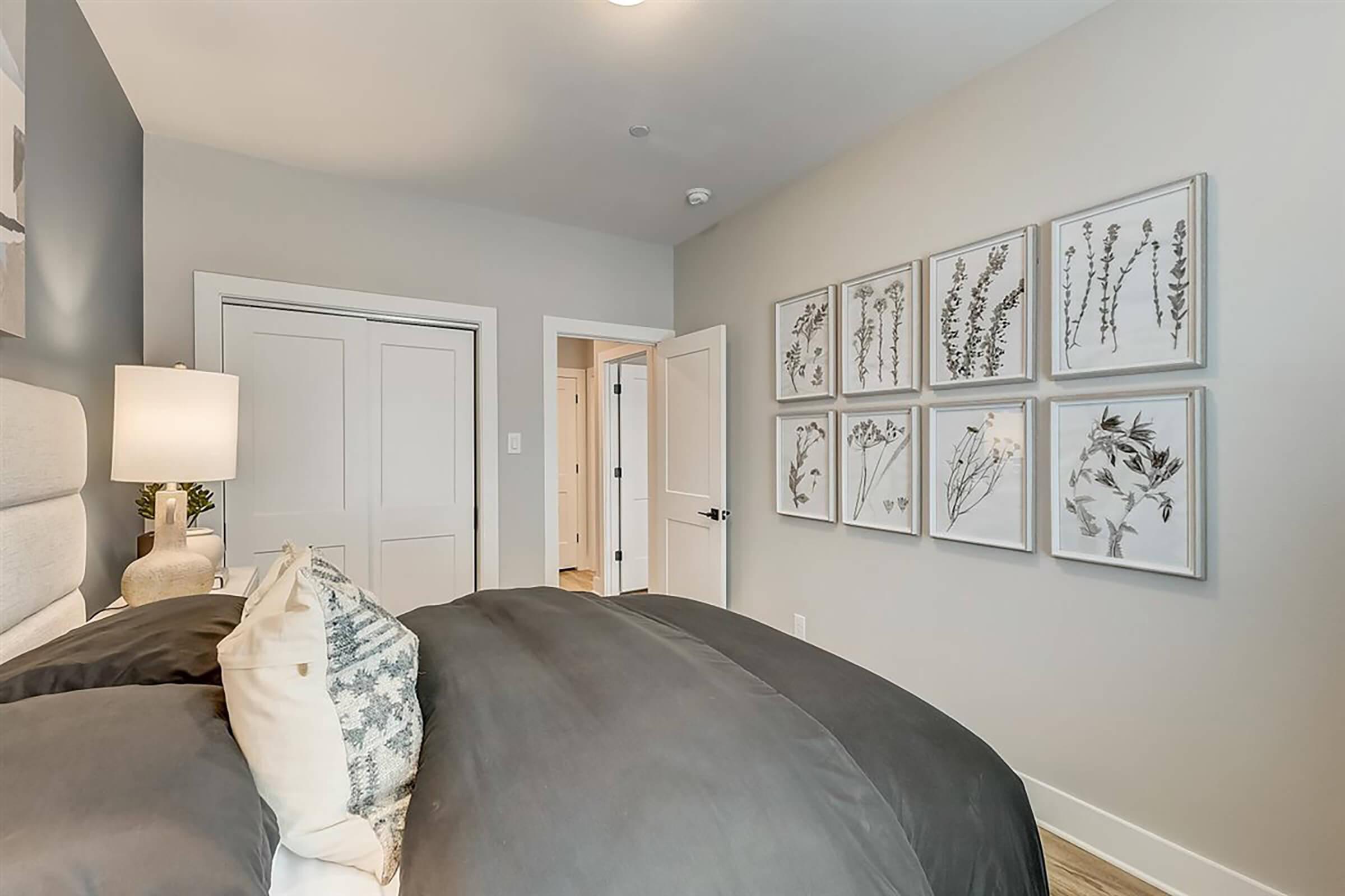 A modern bedroom featuring a gray bedspread with decorative pillows. The walls are painted a light color, and one wall displays a series of framed botanical prints. A closed closet door is visible, and there is natural light coming from the adjacent doorway. The overall decor is minimalistic and contemporary.