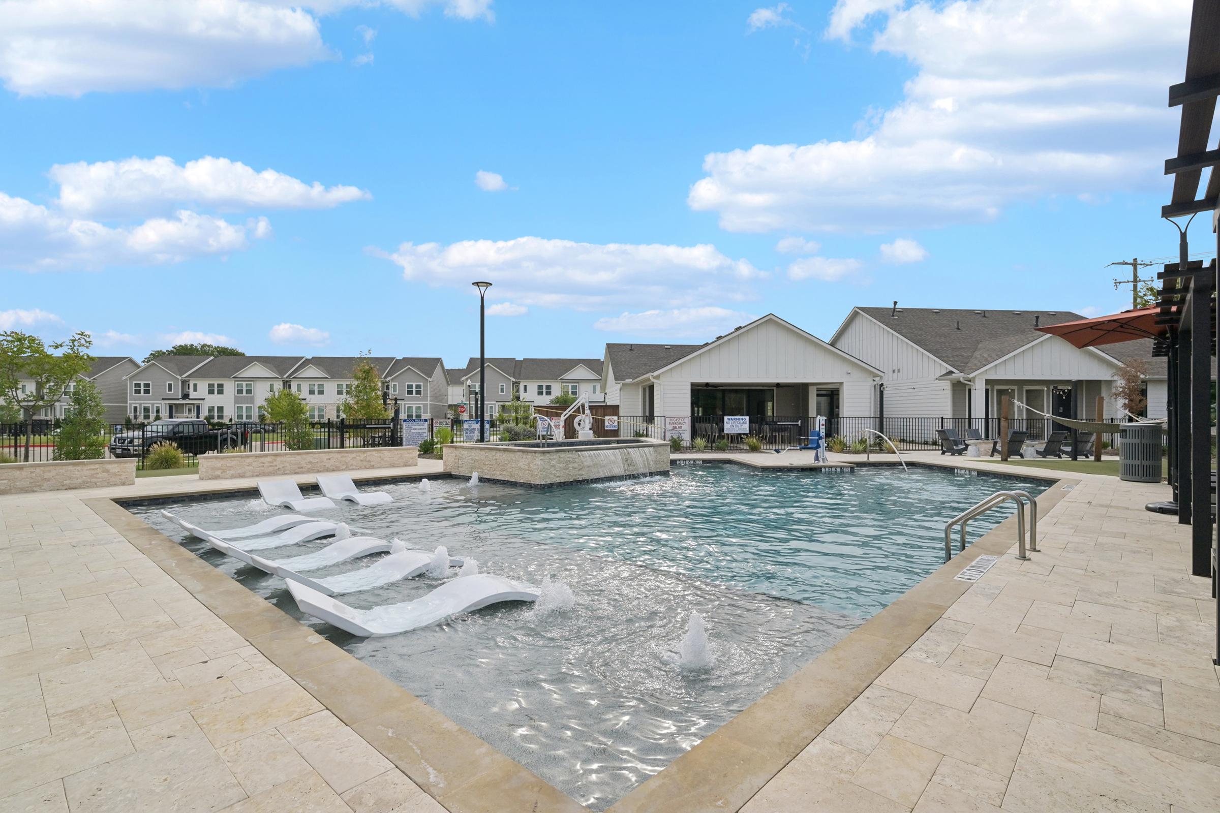 A modern outdoor pool area featuring a shallow swimming pool with fountains, surrounded by lounge chairs. In the background, there are residential buildings with a clear blue sky above and a few clouds. The environment is serene and inviting, perfect for relaxation or leisure activities.