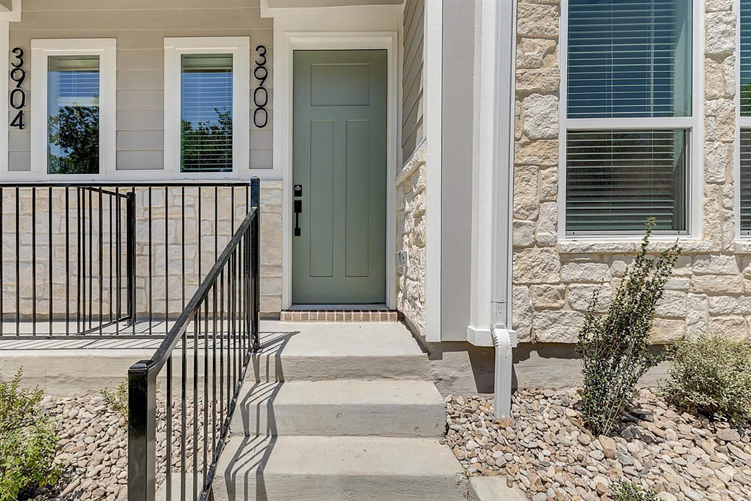 Exterior view of a house entrance featuring a light green front door, stone detailing on the walls, and a small set of concrete steps leading up. A black railing runs alongside the steps, and there are windows on either side of the door. Low landscaping with gravel and small plants is visible.