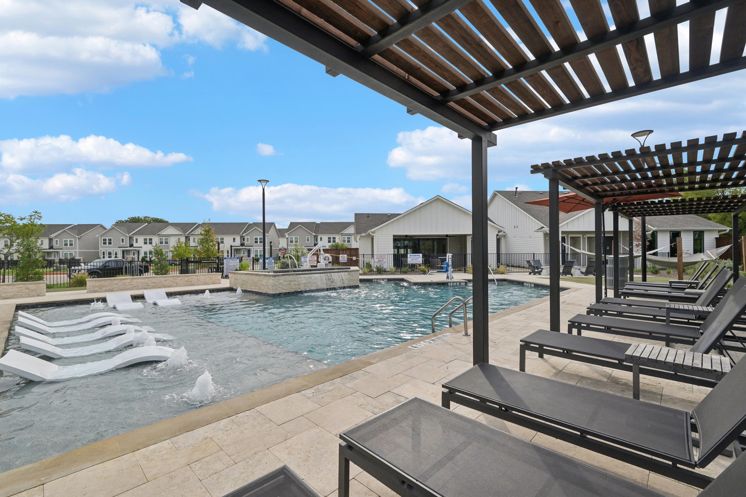 A modern outdoor pool area featuring a spacious swimming pool with water features, surrounded by lounge chairs and shaded pergolas. In the background, there are well-maintained residential buildings and a clear blue sky with some clouds.