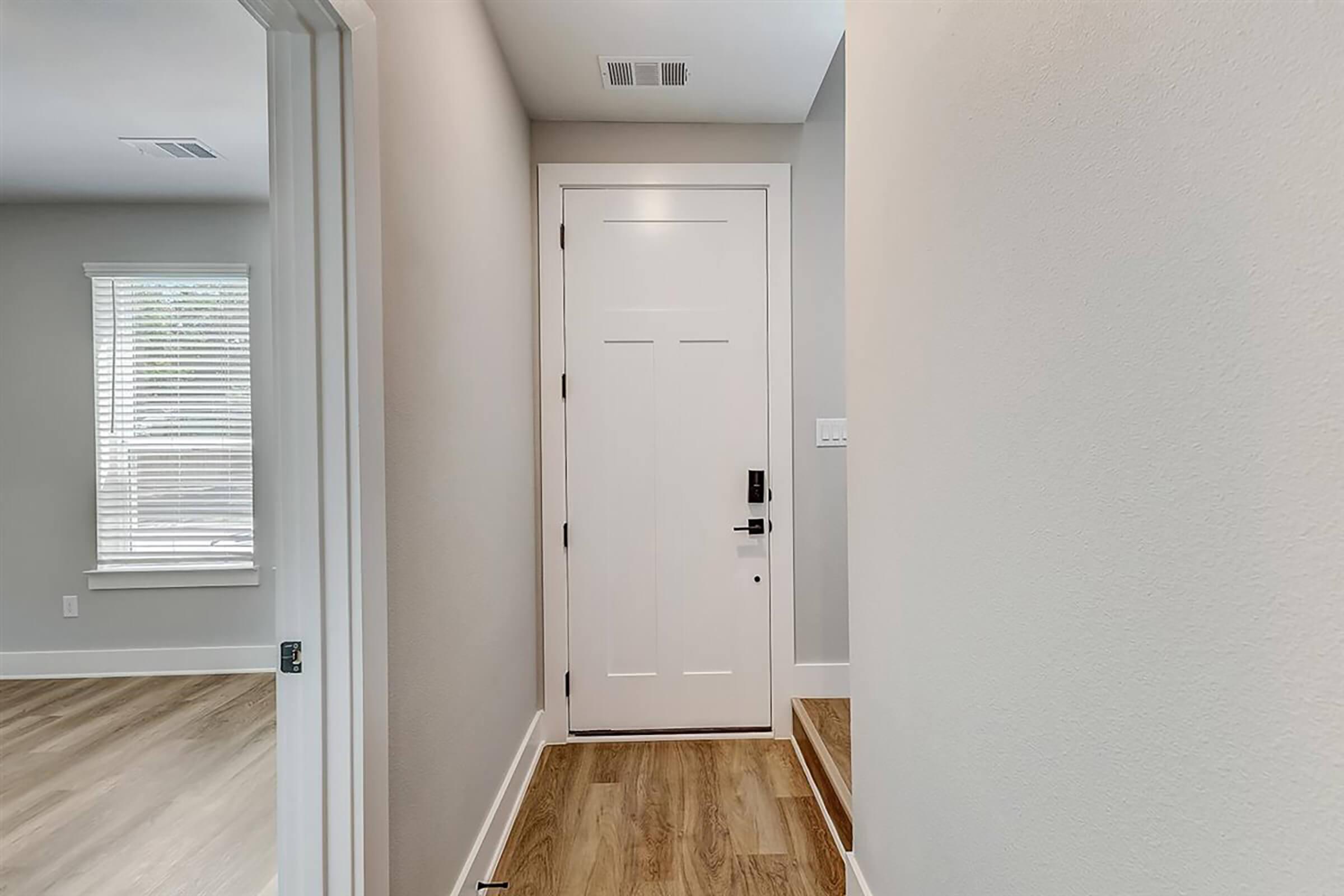 A view of an interior hallway leading to a white front door. The wall is painted light gray, and there are wooden floorboards visible. A set of stairs is located to the right, and a window with blinds can be seen in the background, providing natural light.
