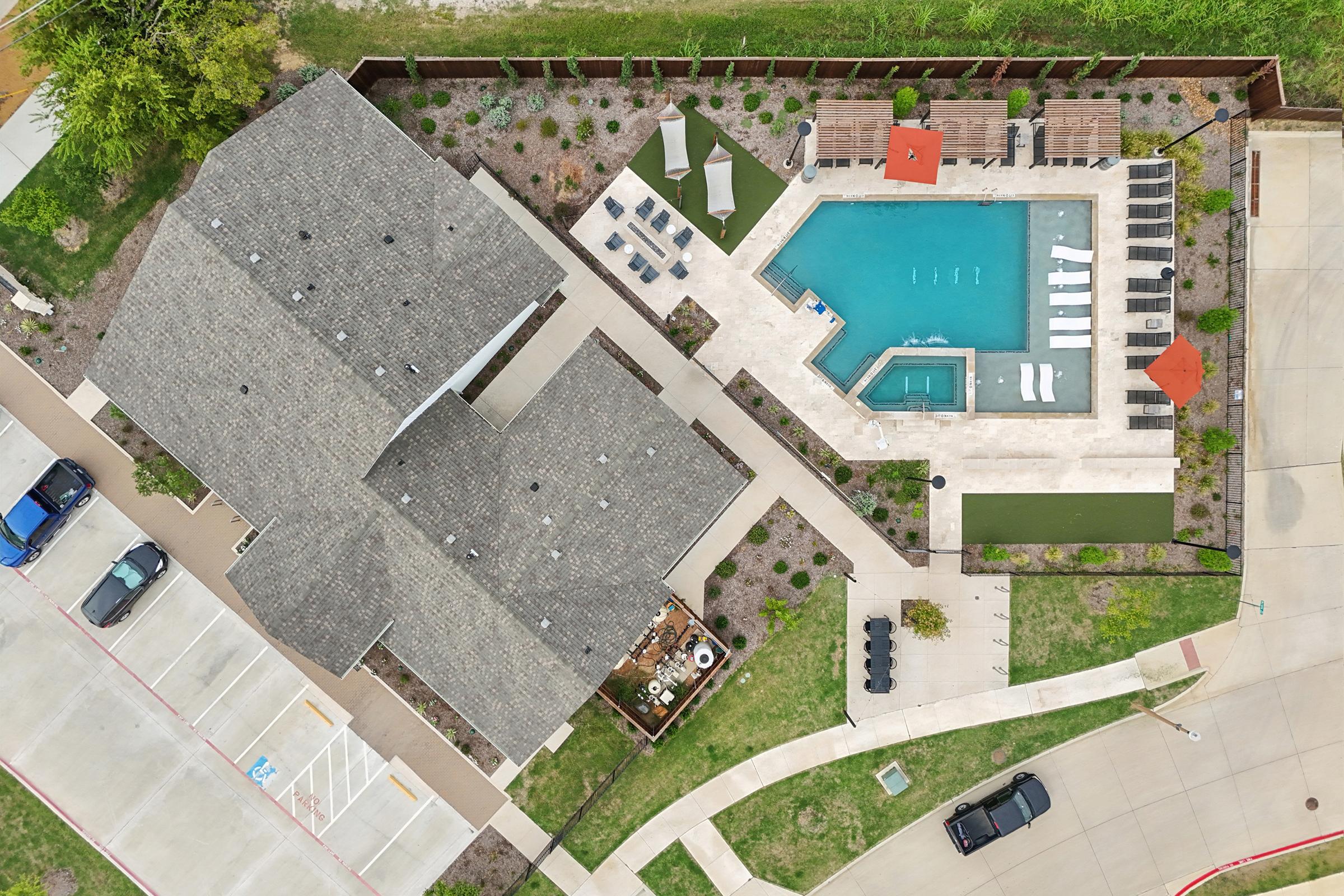 Aerial view of a residential complex featuring a swimming pool with lounge chairs, a hot tub, landscaped gardens, and outdoor seating areas. Nearby, there are parked cars in a paved lot and grassy areas, providing a relaxing atmosphere for residents.