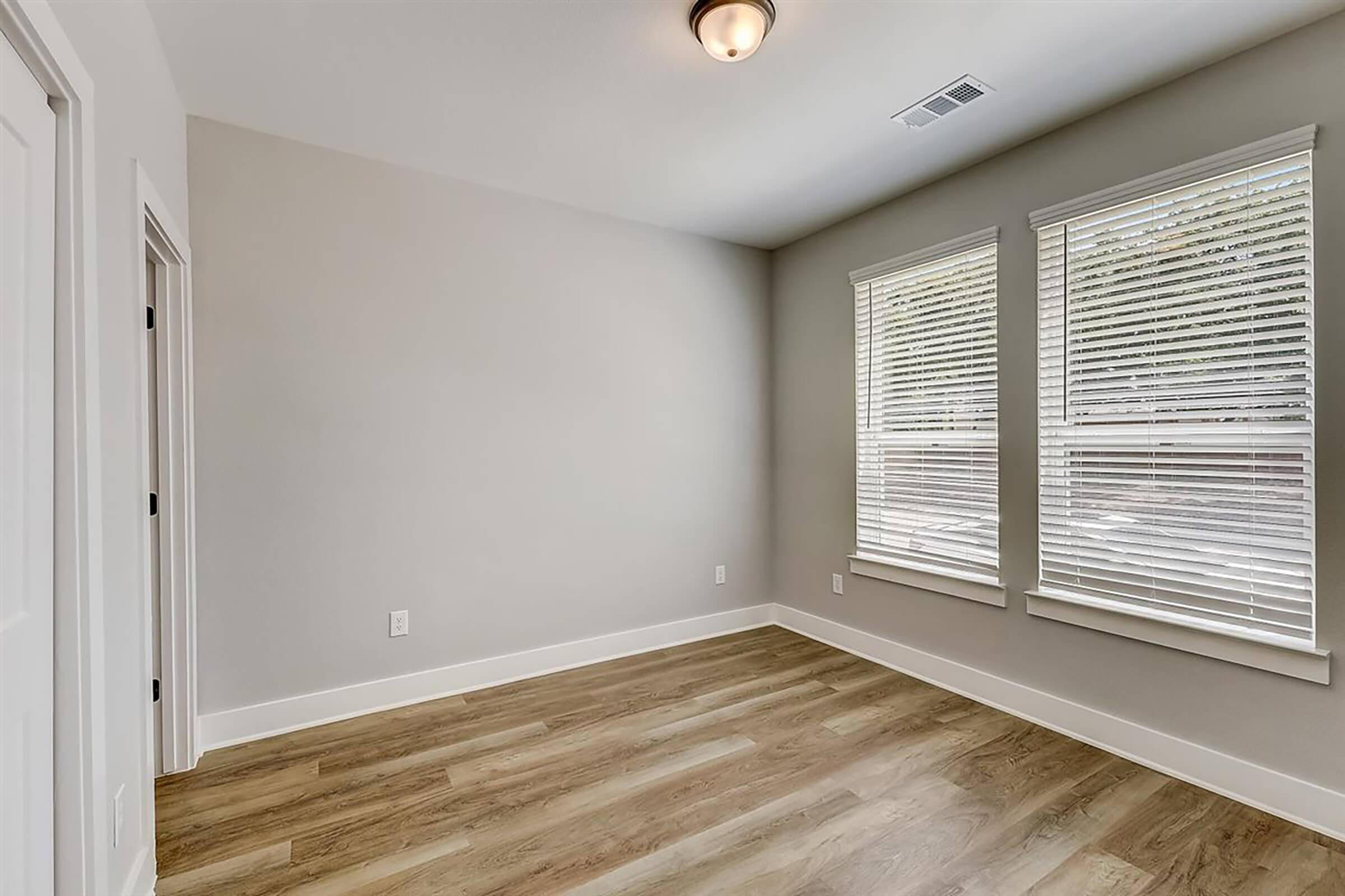 A vacant room featuring light-colored walls and wooden flooring. It has two large windows with blinds, allowing natural light to enter. A ceiling light fixture is installed, and there are two doors leading to different areas. The room appears bright and spacious, suitable for various uses.