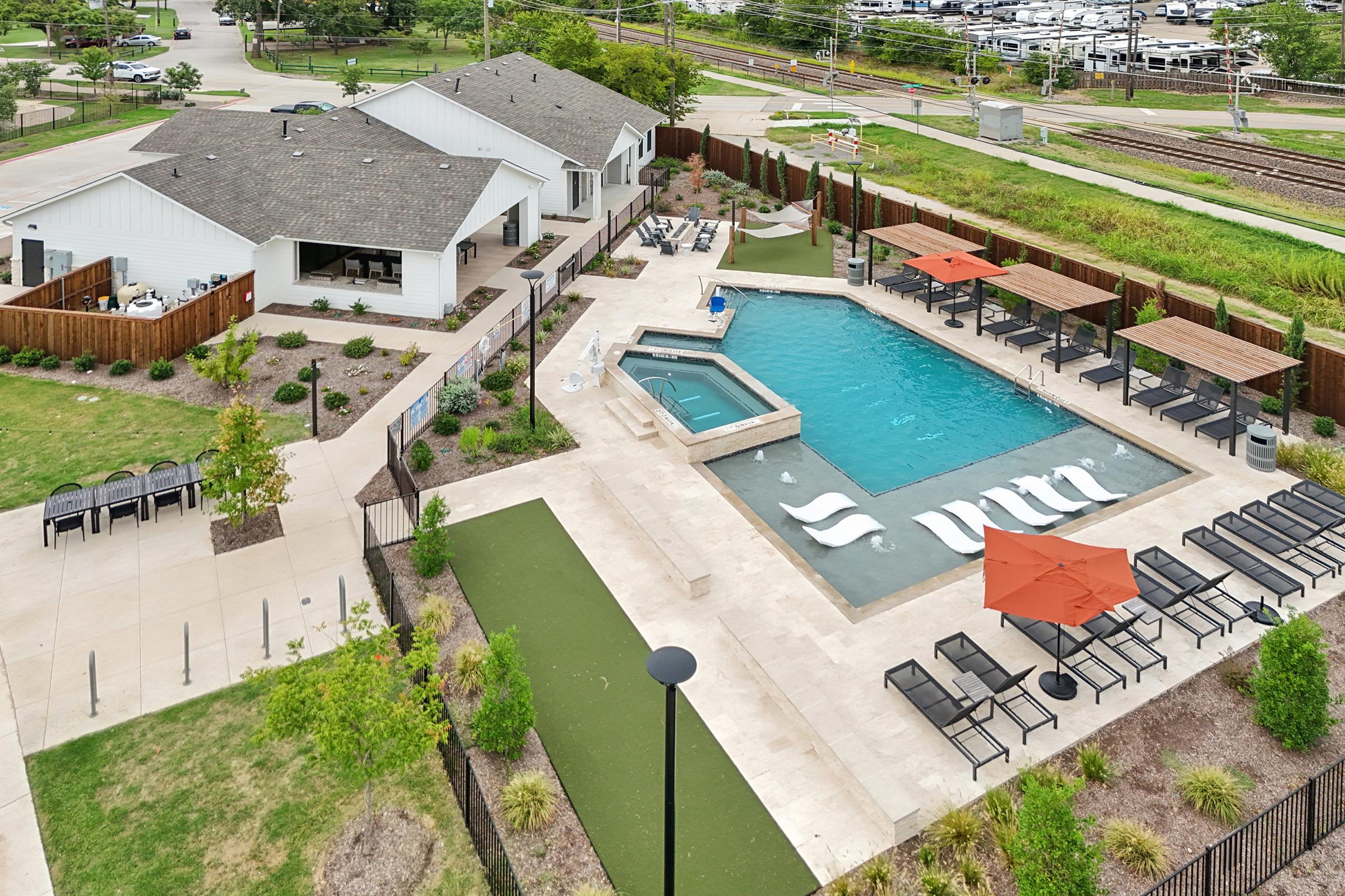 Aerial view of a community pool area featuring a large swimming pool with lounge chairs and umbrellas, surrounded by landscaped greenery. Nearby, there are several seating areas and a hot tub. In the background, there are residential buildings and a parking area, along with a train track visible.